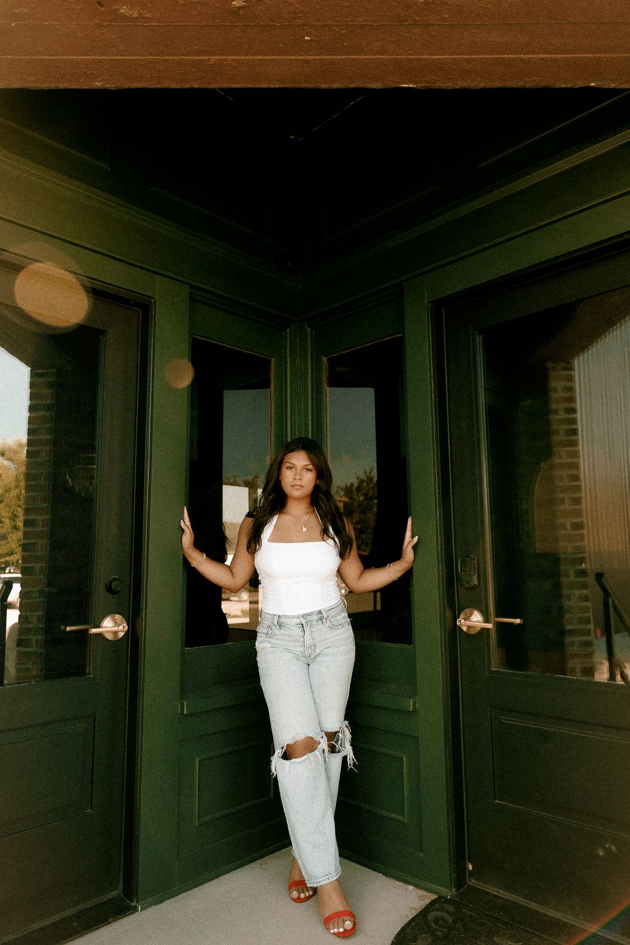 Young woman in white crop top and ripped jeans poses between green double doors of a brick building.