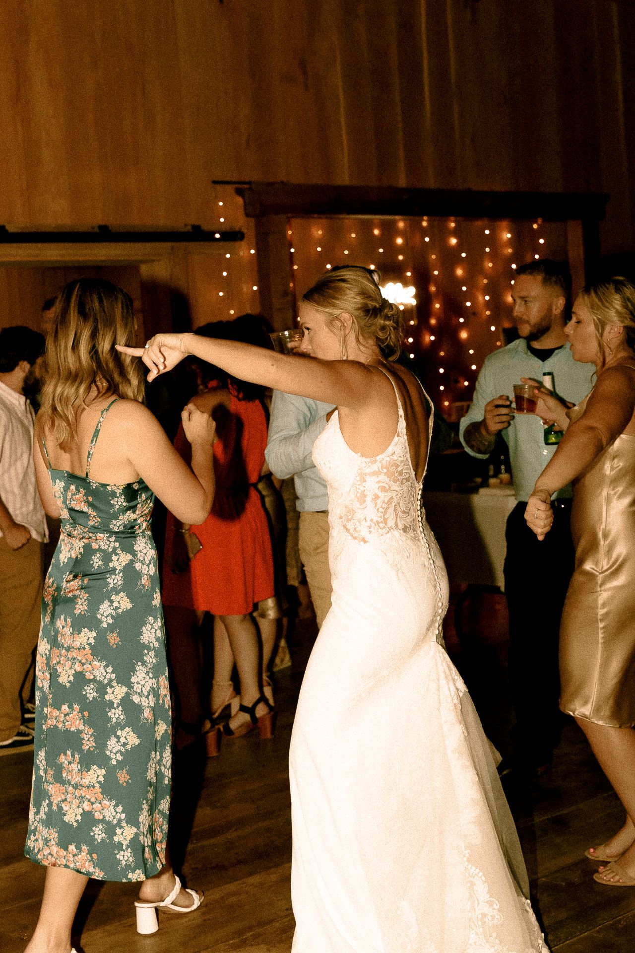Bride in white lace gown dancing with guests at a rustic wedding reception with warm string lights backdrop.