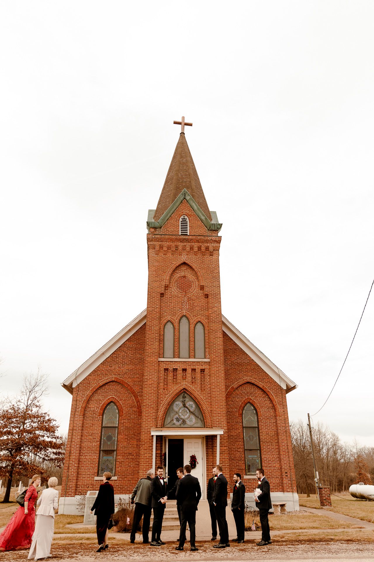 Wedding party gathered outside a historic red brick church with a steeple and cross on an overcast day.