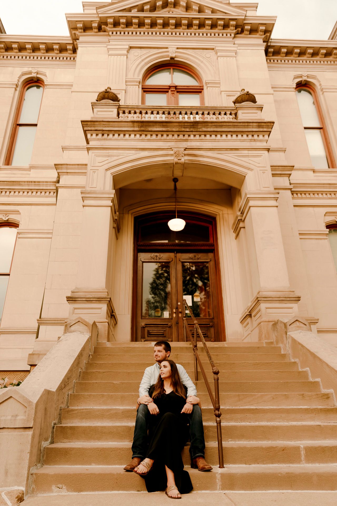 Couple sitting on steps of historic building with ornate architecture, sharing a romantic moment during engagement photo session.
