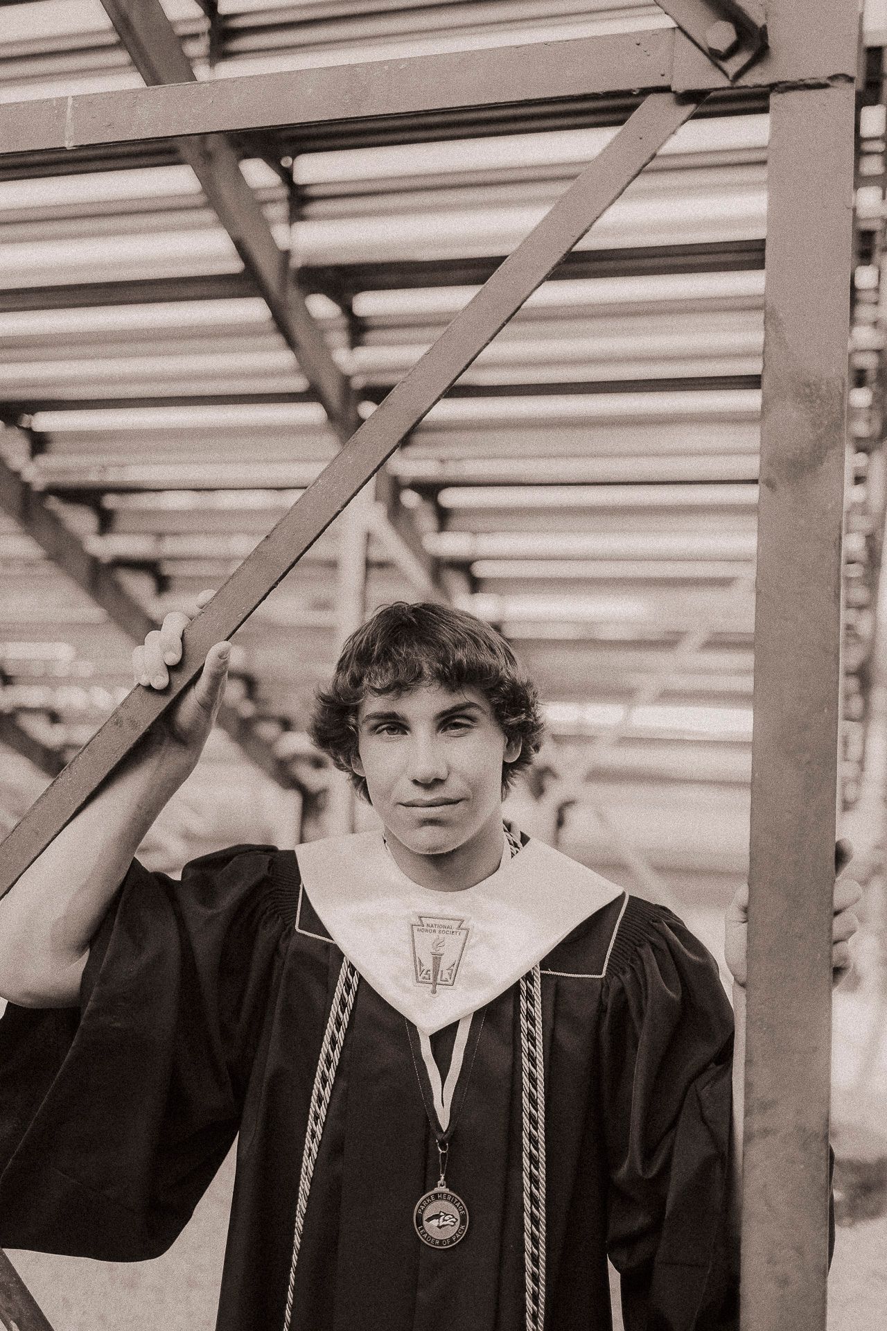 Young man in graduation gown and medal poses by metal beams in sepia-toned senior portrait photo.