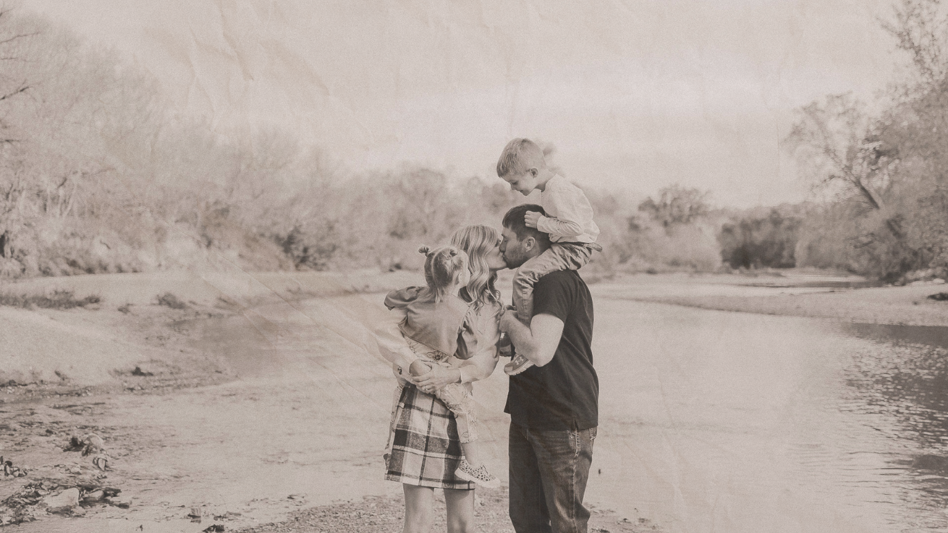 Black and white photo of a father with children by a river, one child on his shoulders, outdoors.