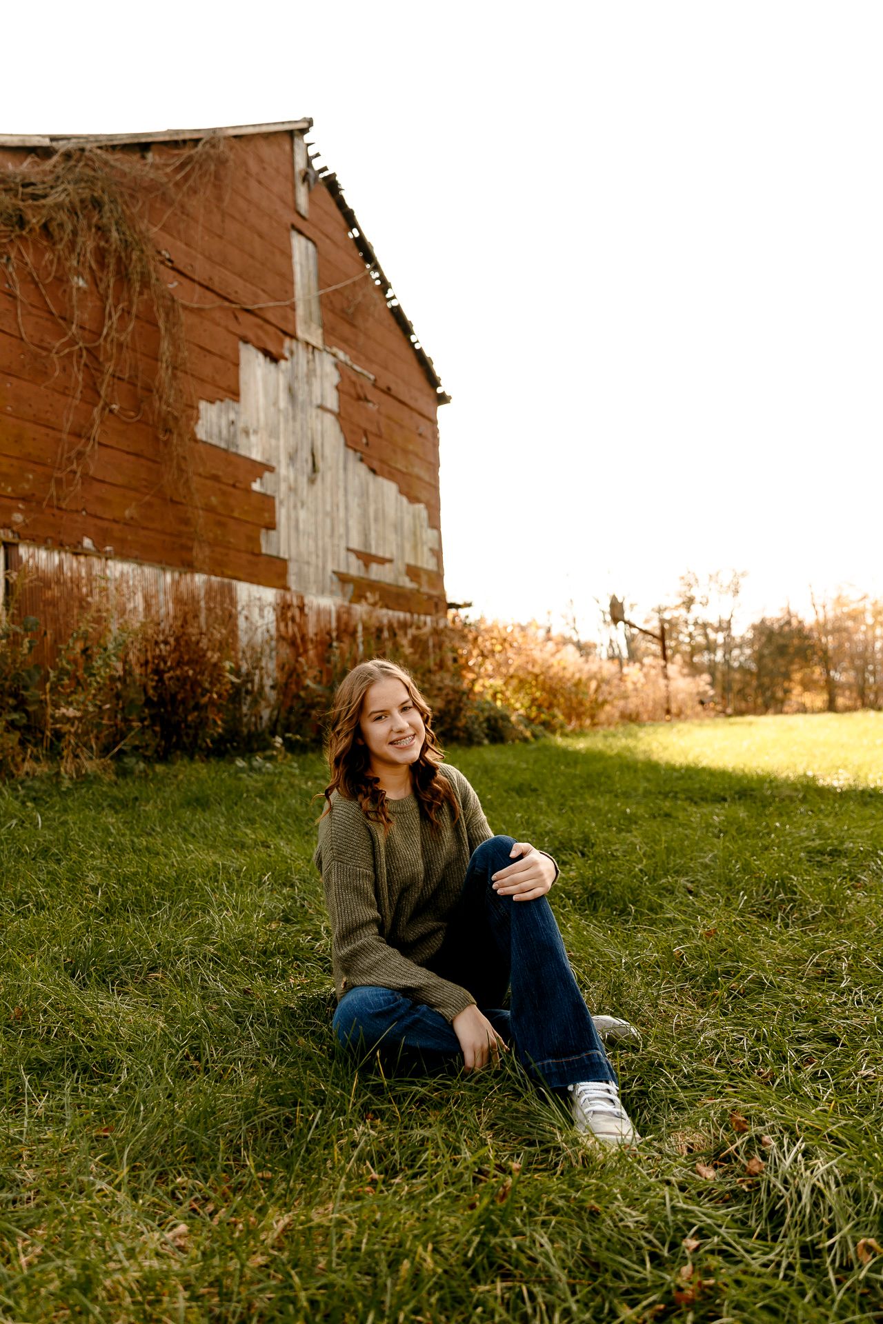 Young woman in olive sweater and jeans sits smiling on grass beside a rustic red barn in golden autumn light.