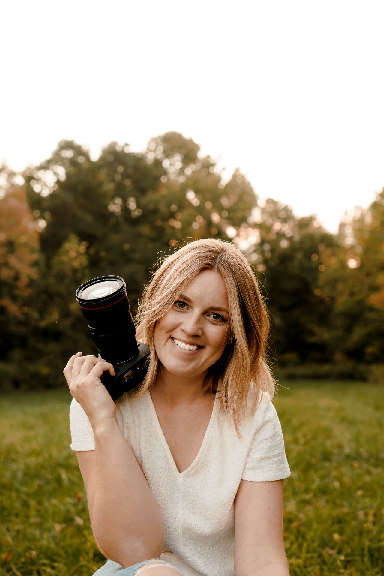 Smiling blonde woman holding a DSLR camera outdoors in a green park setting during golden hour.