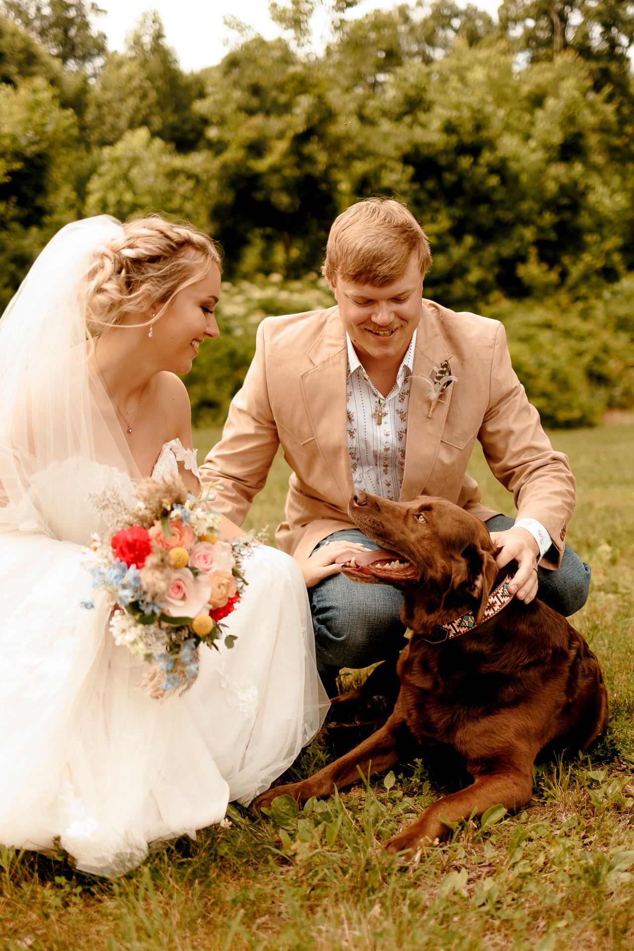 Bride and groom in wedding attire petting a chocolate Labrador dog outdoors on green grass with trees in background.