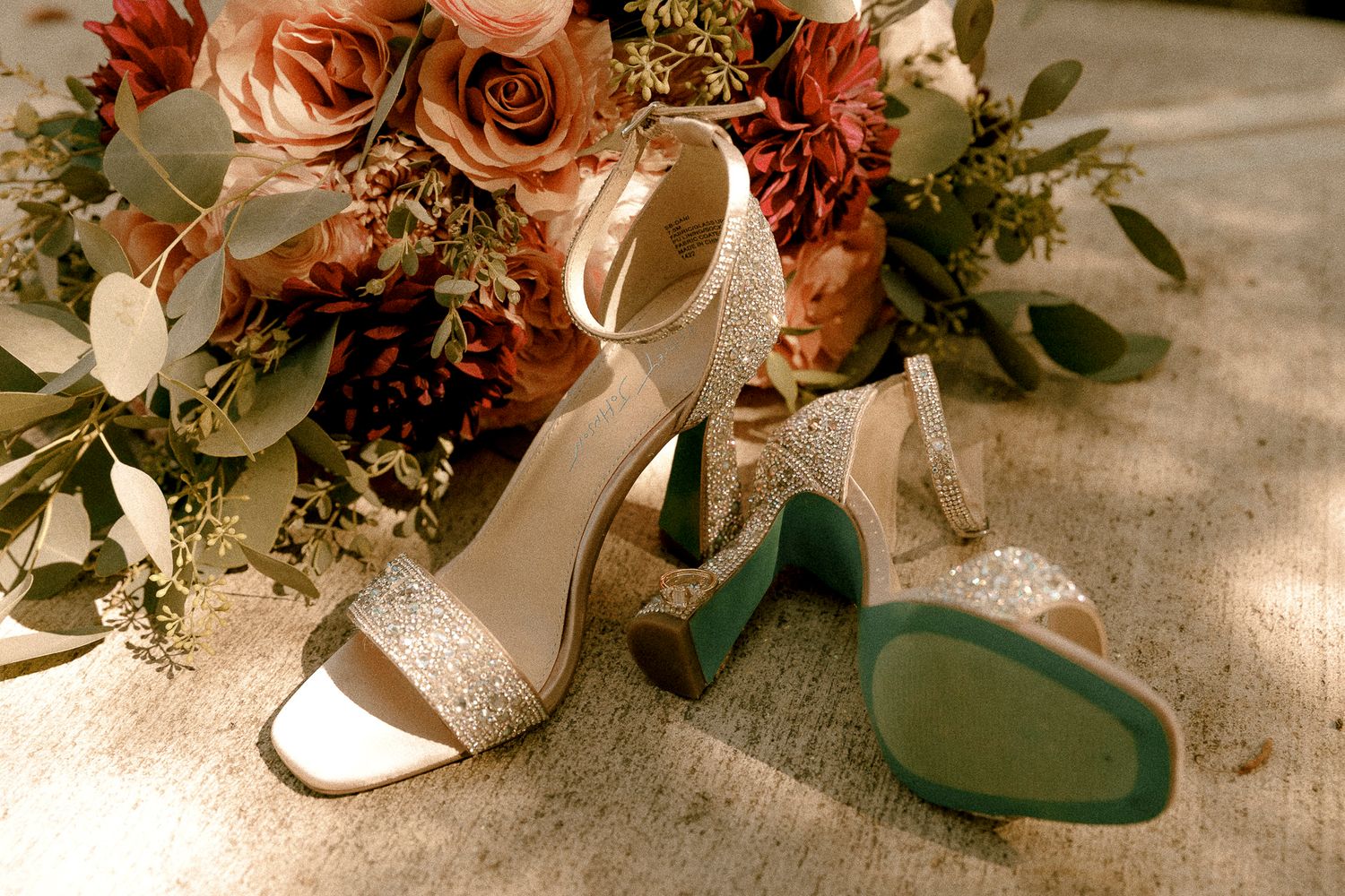 Glittery bridal heels with teal soles beside a lush bouquet of pink and red roses on a textured surface.