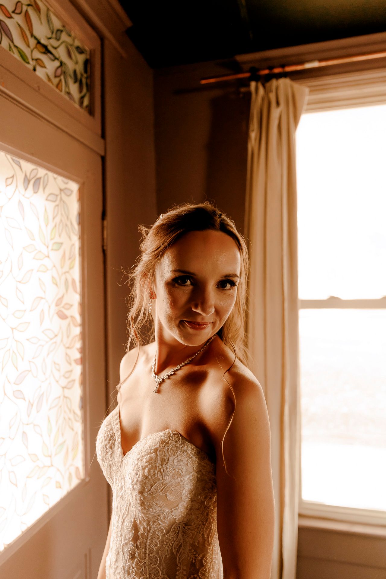 Bride in strapless lace wedding gown smiling softly near sunlit windows with decorative glass panels.