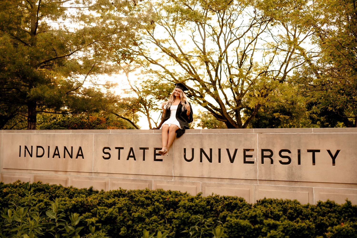 Graduate poses on Indiana State University entrance sign surrounded by lush green trees at golden hour.