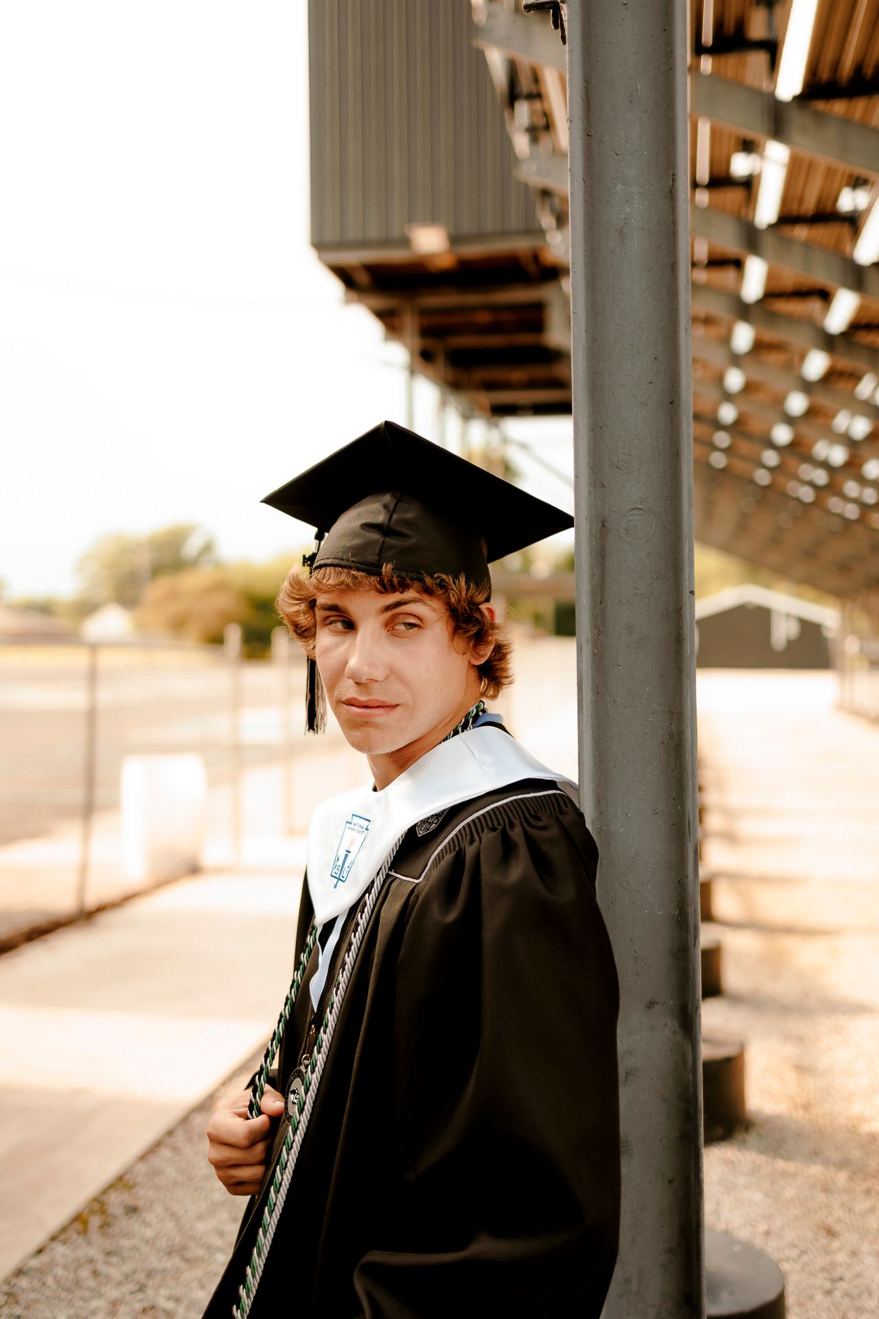 Young man in black graduation cap and gown poses confidently near a metal pole at an outdoor venue.
