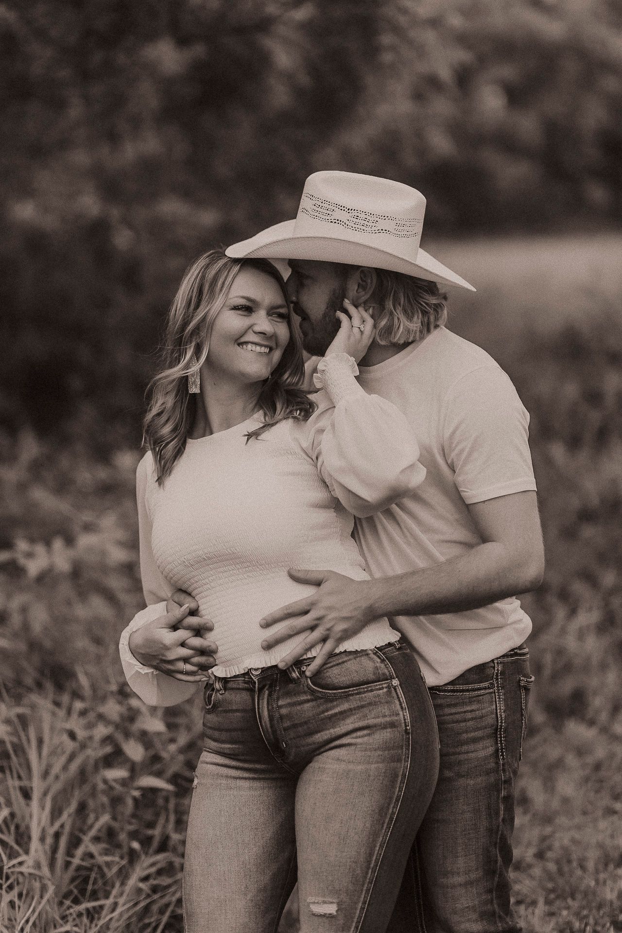 Sepia-toned photo of a couple embracing outdoors, man in cowboy hat kissing womans cheek while holding her bump.