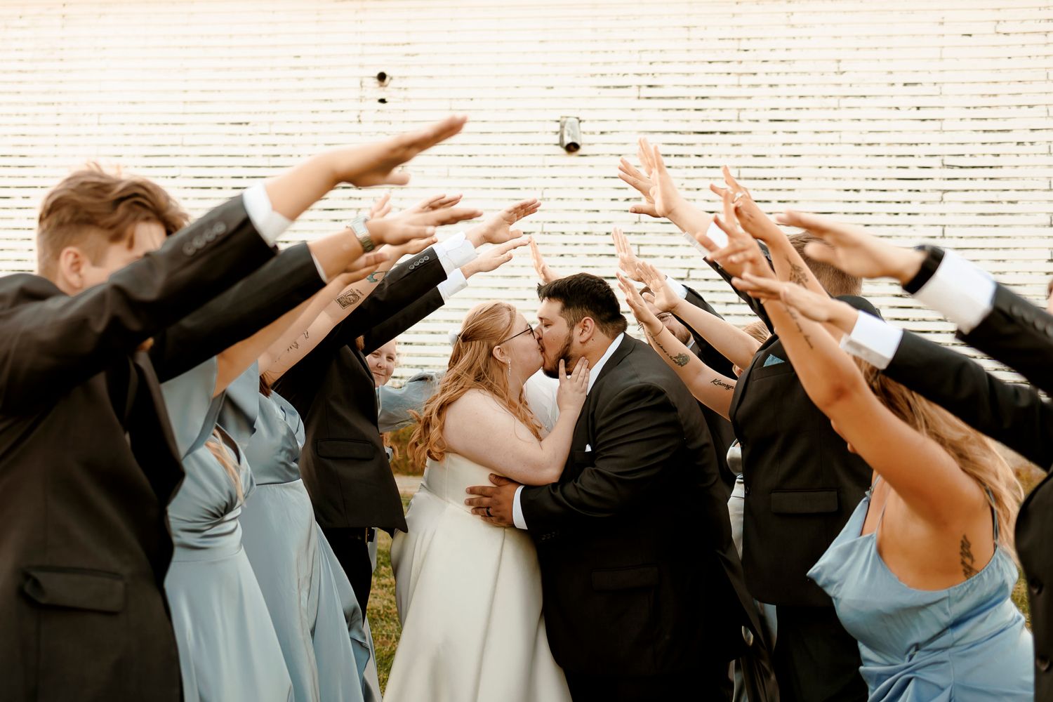 Wedding couple kissing under arch of raised hands from bridal party in blue dresses and dark suits outdoors.