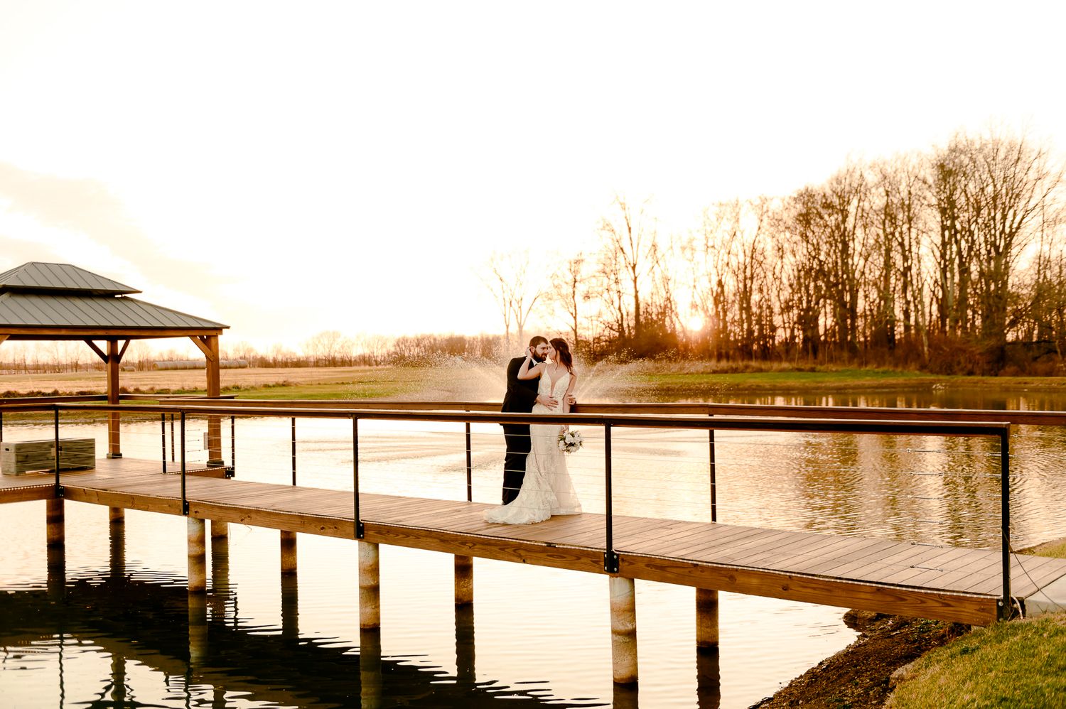 Bride and groom kissing on a wooden dock over a serene pond at golden hour with trees in the background.