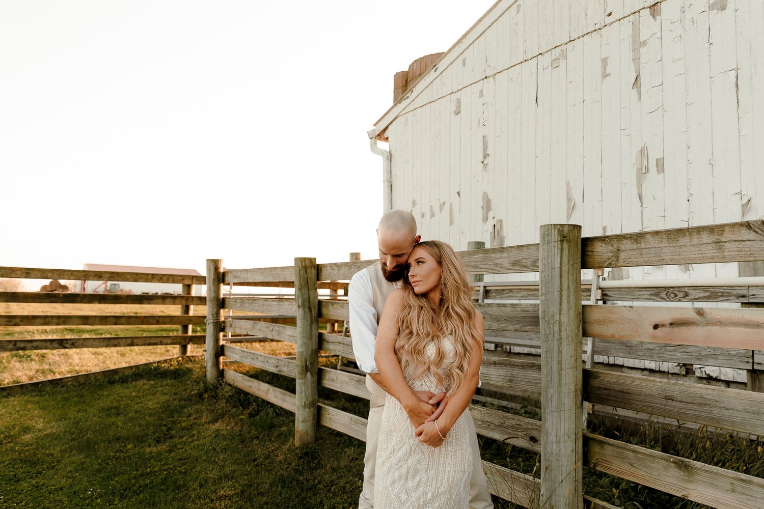 Couple embracing by a rustic wooden fence near a white barn at golden hour in a romantic outdoor setting.