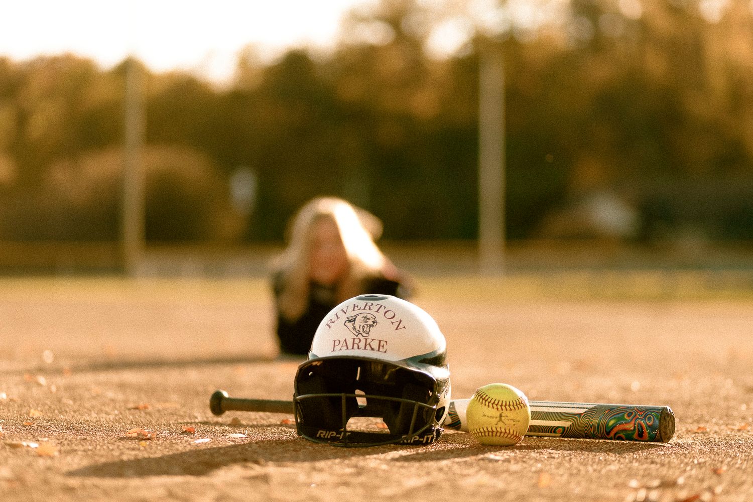 Softball helmet and bat on dirt field with blurred player in background during golden hour.