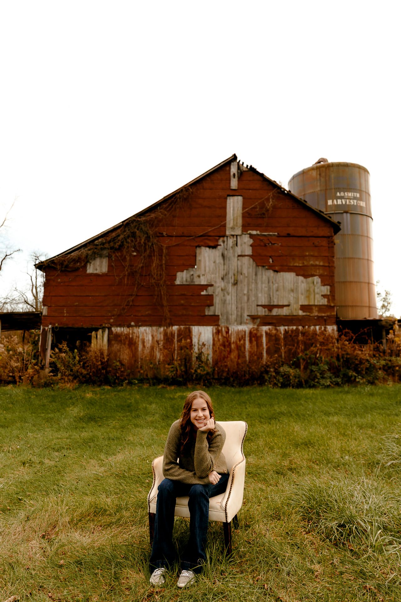 Young woman sitting in a white armchair on grass in front of a rustic red barn and grain silo.