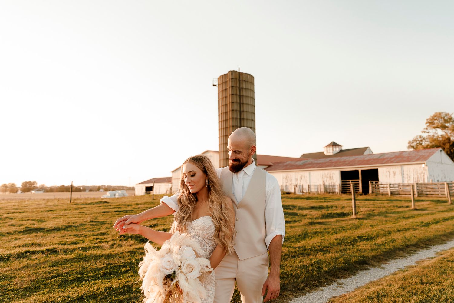 Bride and groom walking on farm path at golden hour, white barn and silo in background.