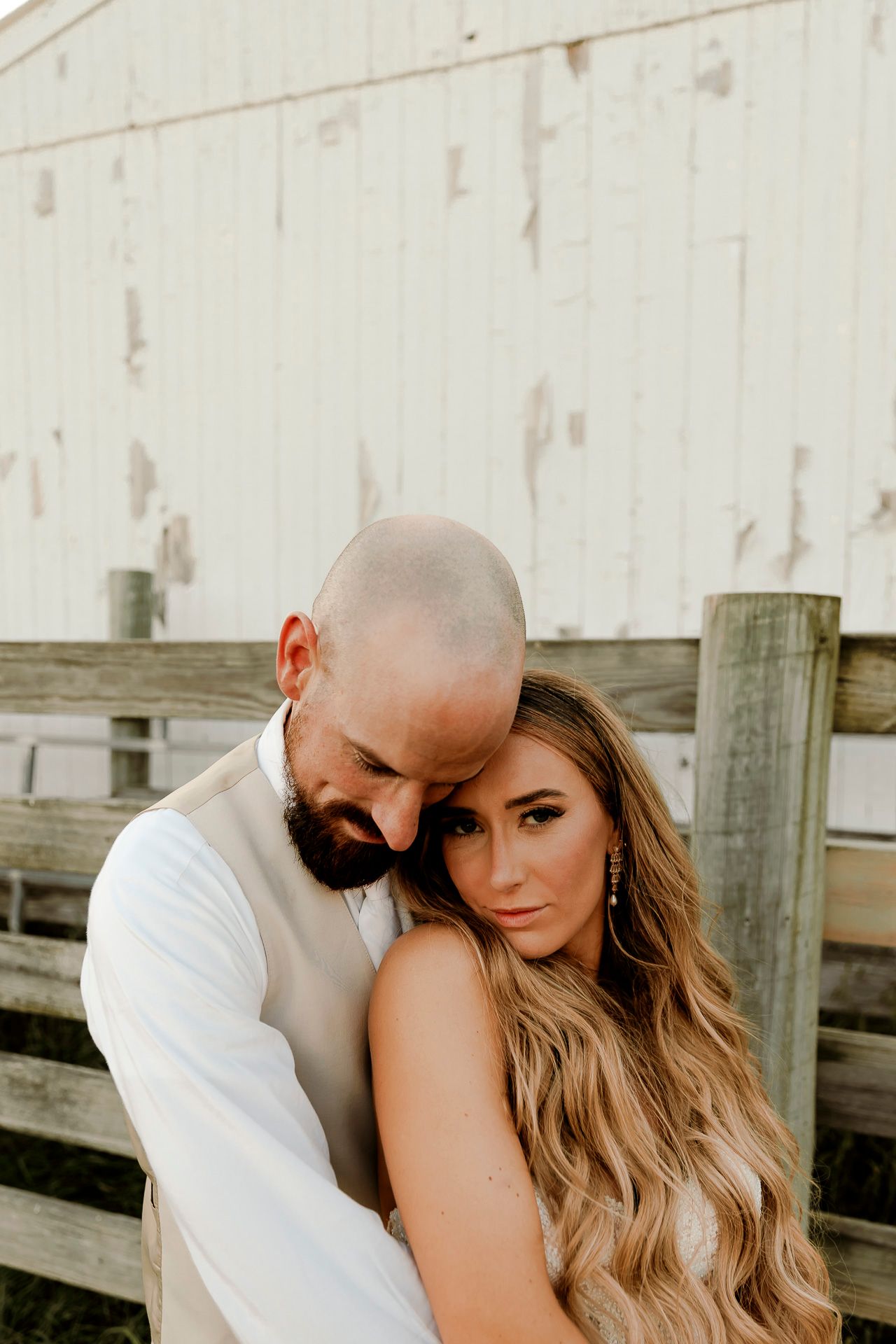 A couple embraces romantically near a rustic wooden fence and white barn, the man resting his head on the woman's hair.