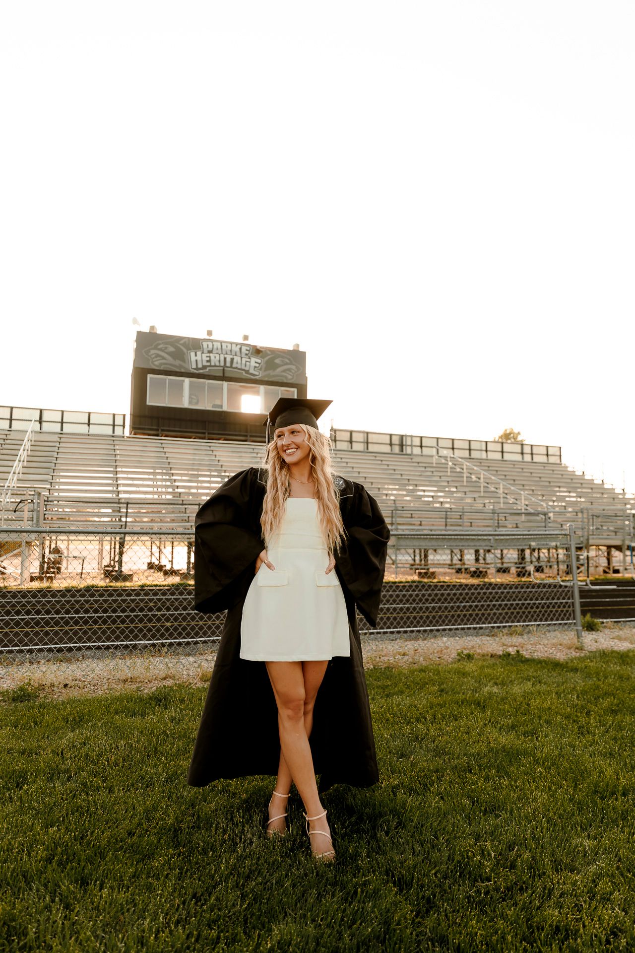 Smiling graduate in black cap and gown over white dress poses on green grass in front of stadium bleachers.
