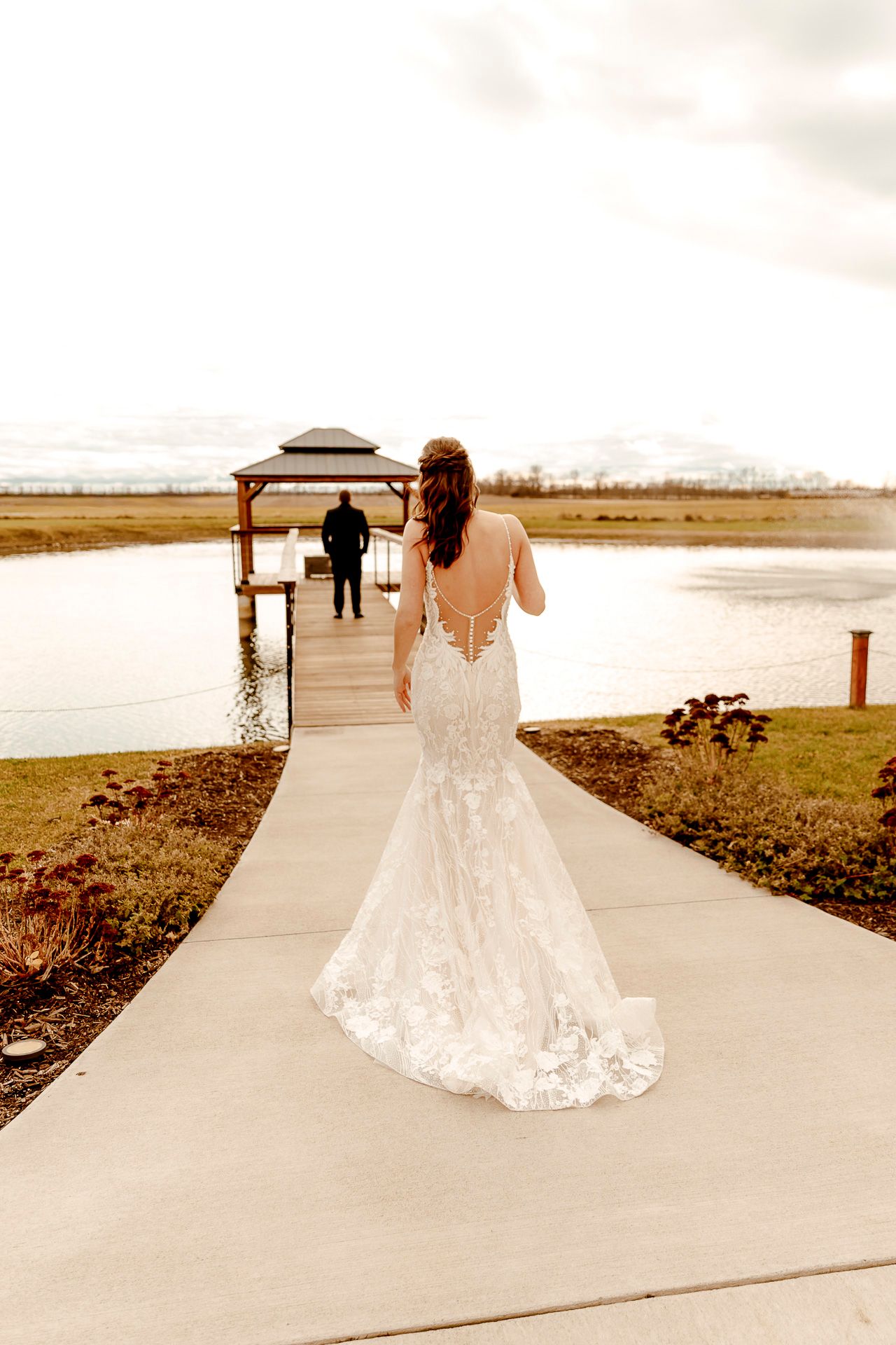 A bride in a lace mermaid wedding dress walks down a concrete path toward a gazebo by a lake.