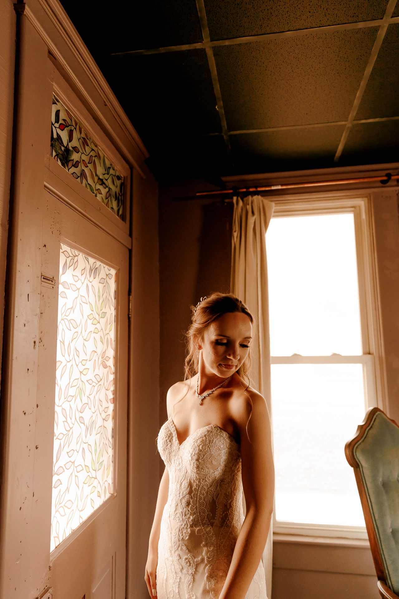 Bride in a strapless lace wedding gown stands by a sunlit window in a vintage room, looking down gracefully.