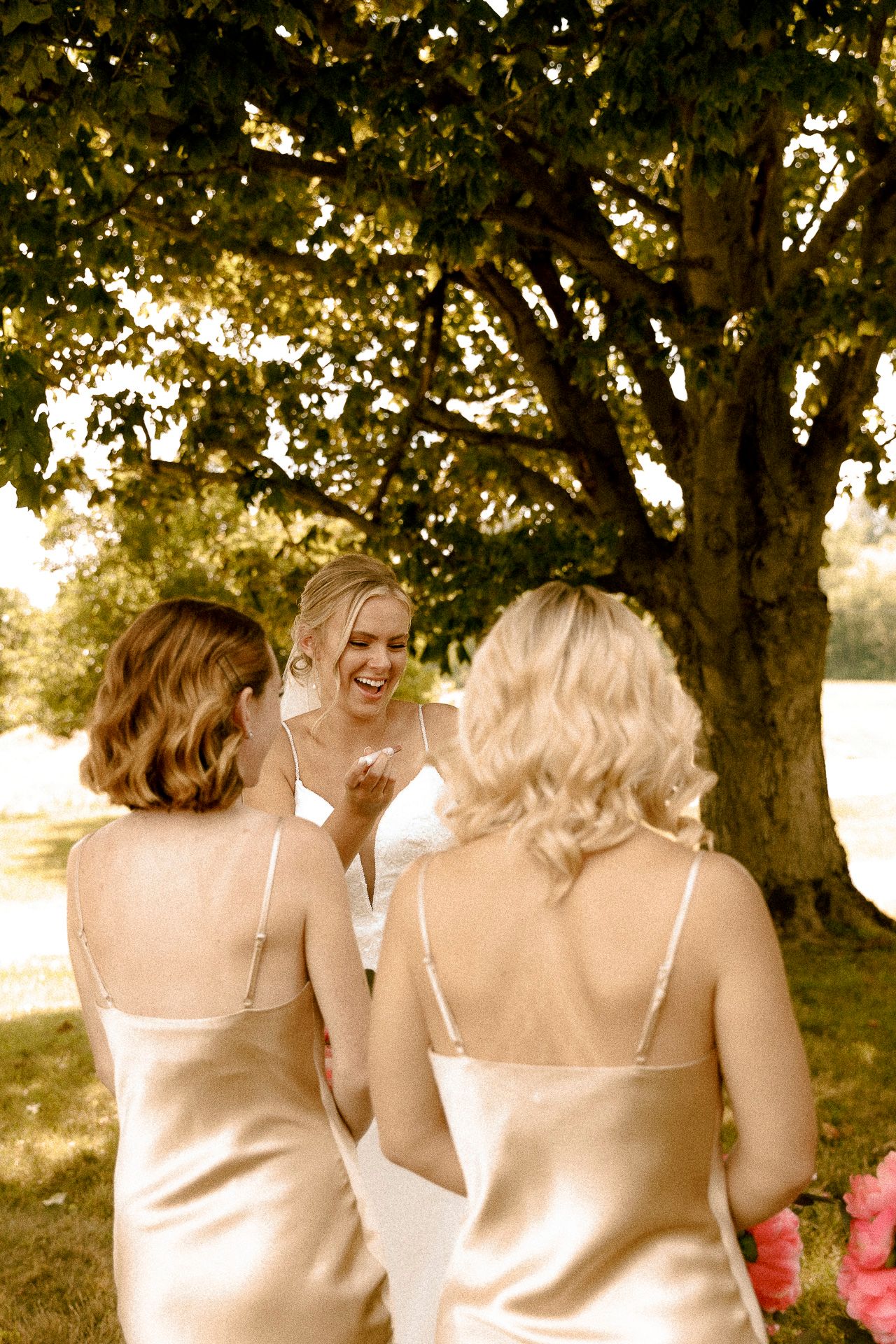 Bride in white dress laughing with two bridesmaids in gold satin gowns under a large tree outdoors.