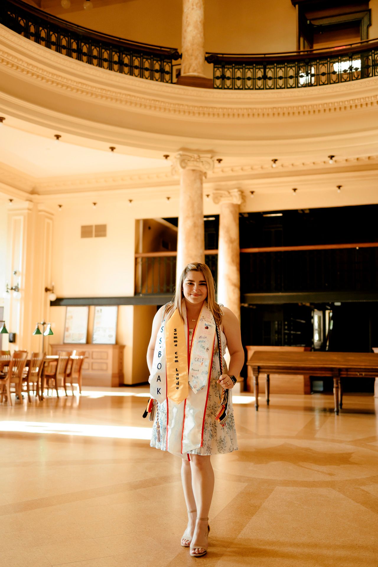 Young woman in graduation stole poses in elegant rotunda building with marble columns and warm lighting.