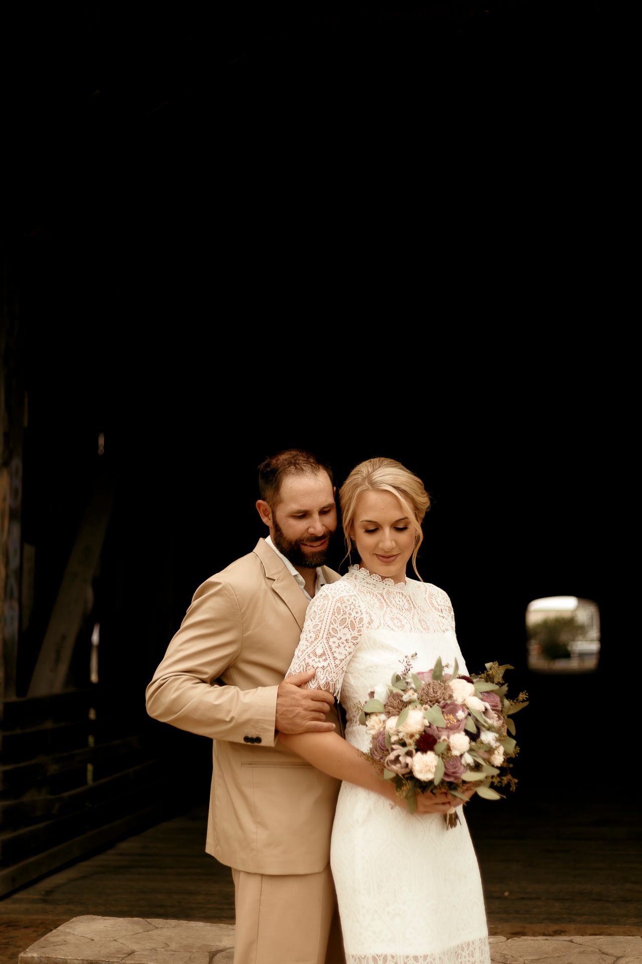 Bride and groom embrace in a covered bridge, bride holding a bouquet of pink and white flowers in a lace wedding dress.