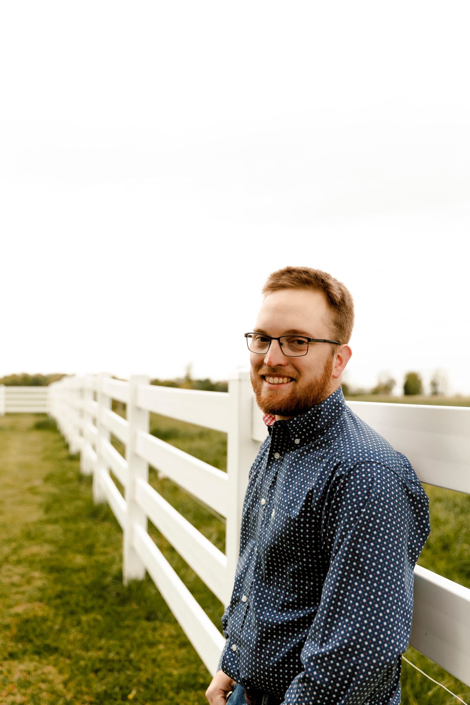 Smiling bearded man with glasses in a navy polka dot shirt leaning against a white wooden fence in a green field.
