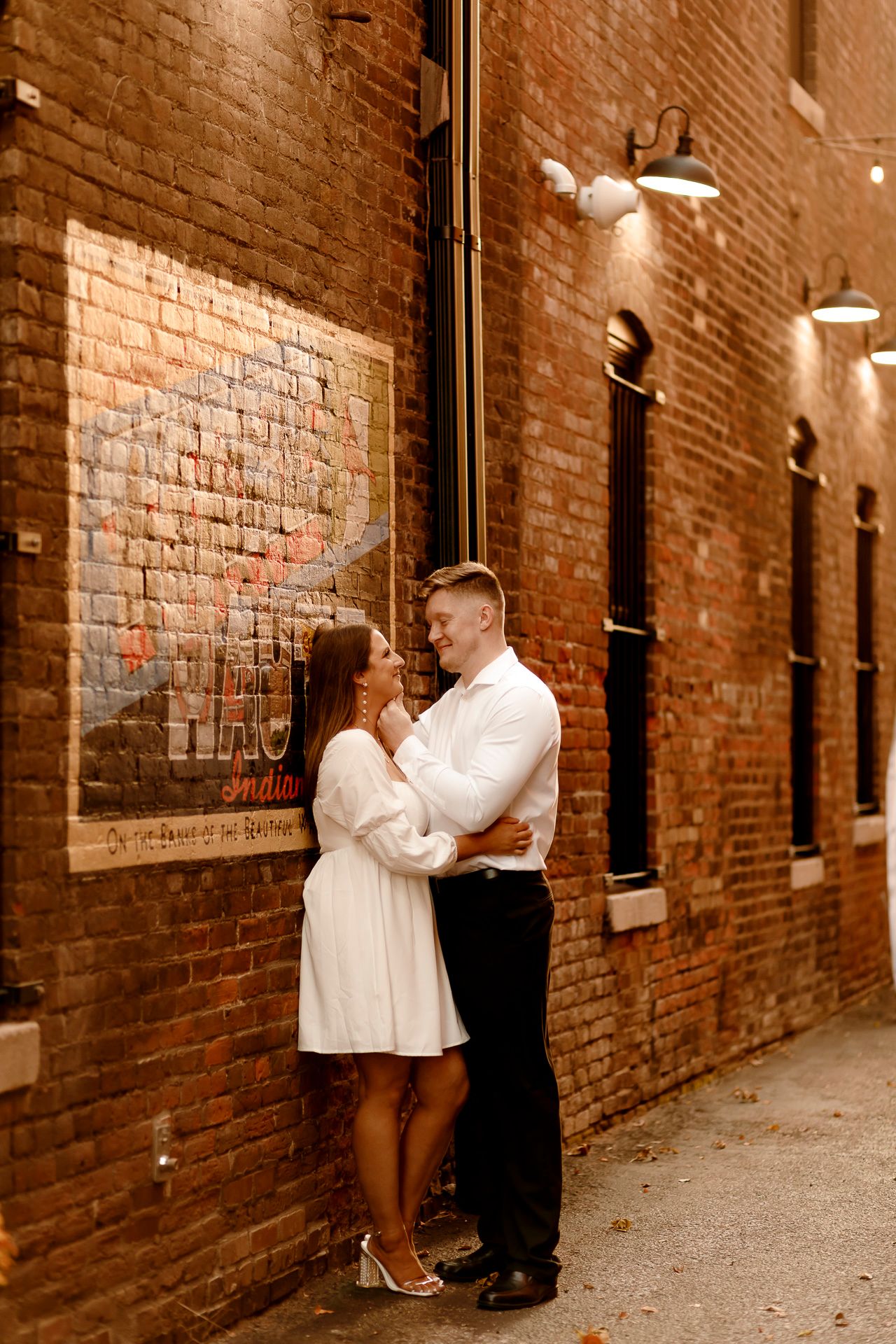 Couple embracing against a rustic brick wall with vintage signage and warm lights in a charming urban alley.