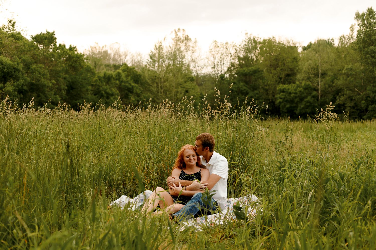 Couple sitting together in tall green grass field, sharing a tender moment during an outdoor engagement photo session.