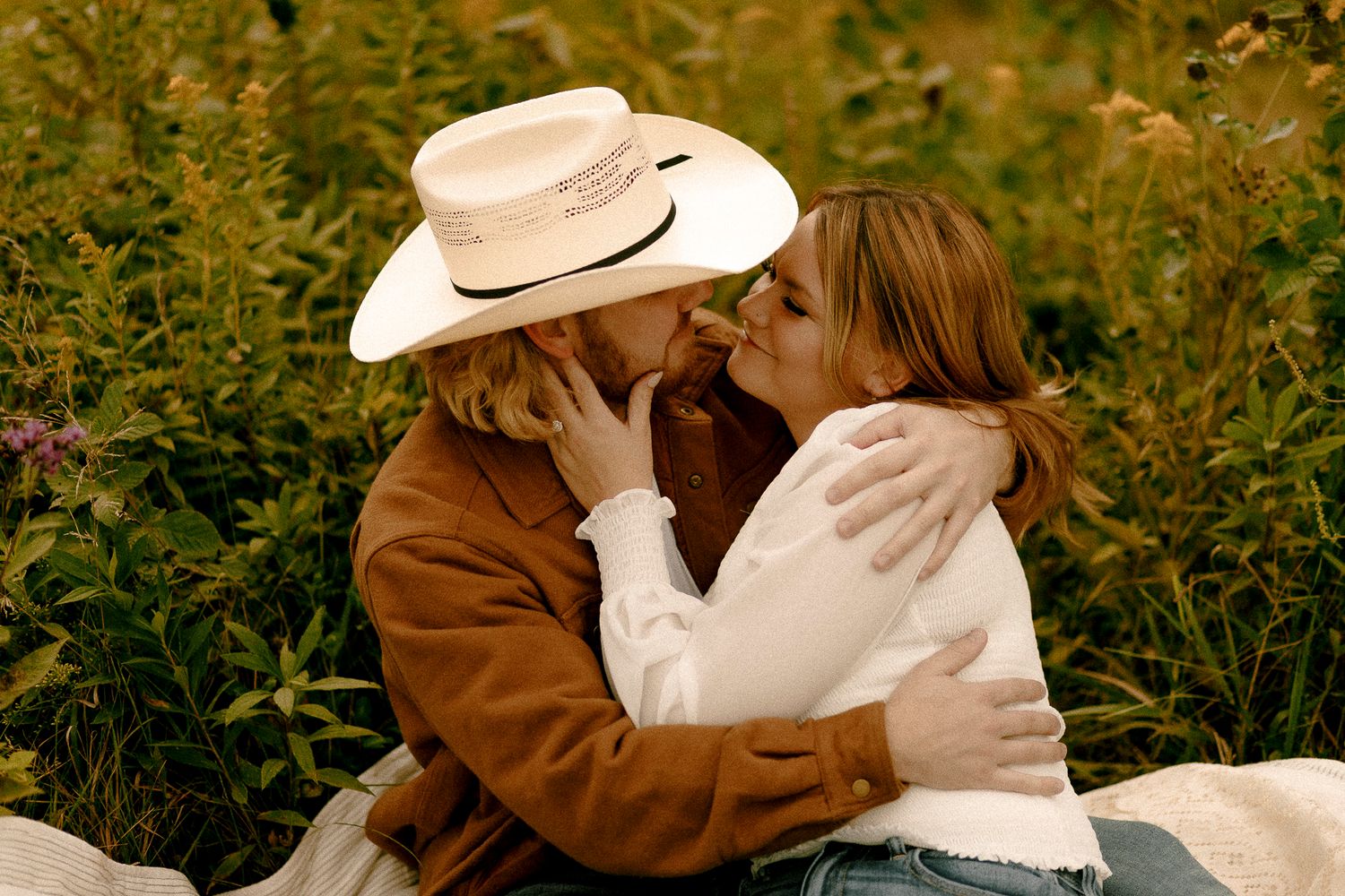 Couple sharing a romantic kiss in a wildflower field, man wearing a white cowboy hat and brown jacket.