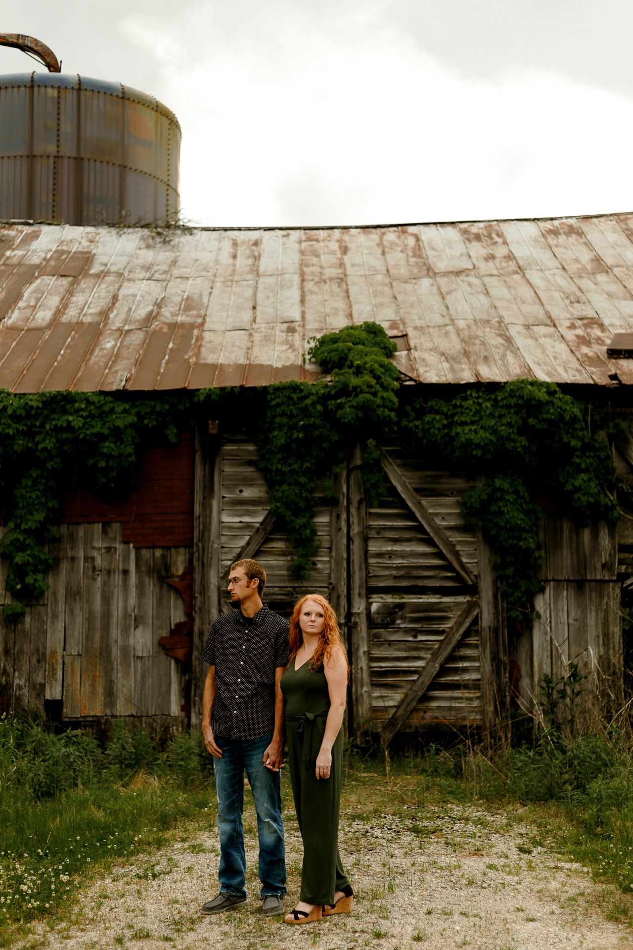 A couple stands hand in hand before a rustic weathered barn with a silo, surrounded by lush green vines.