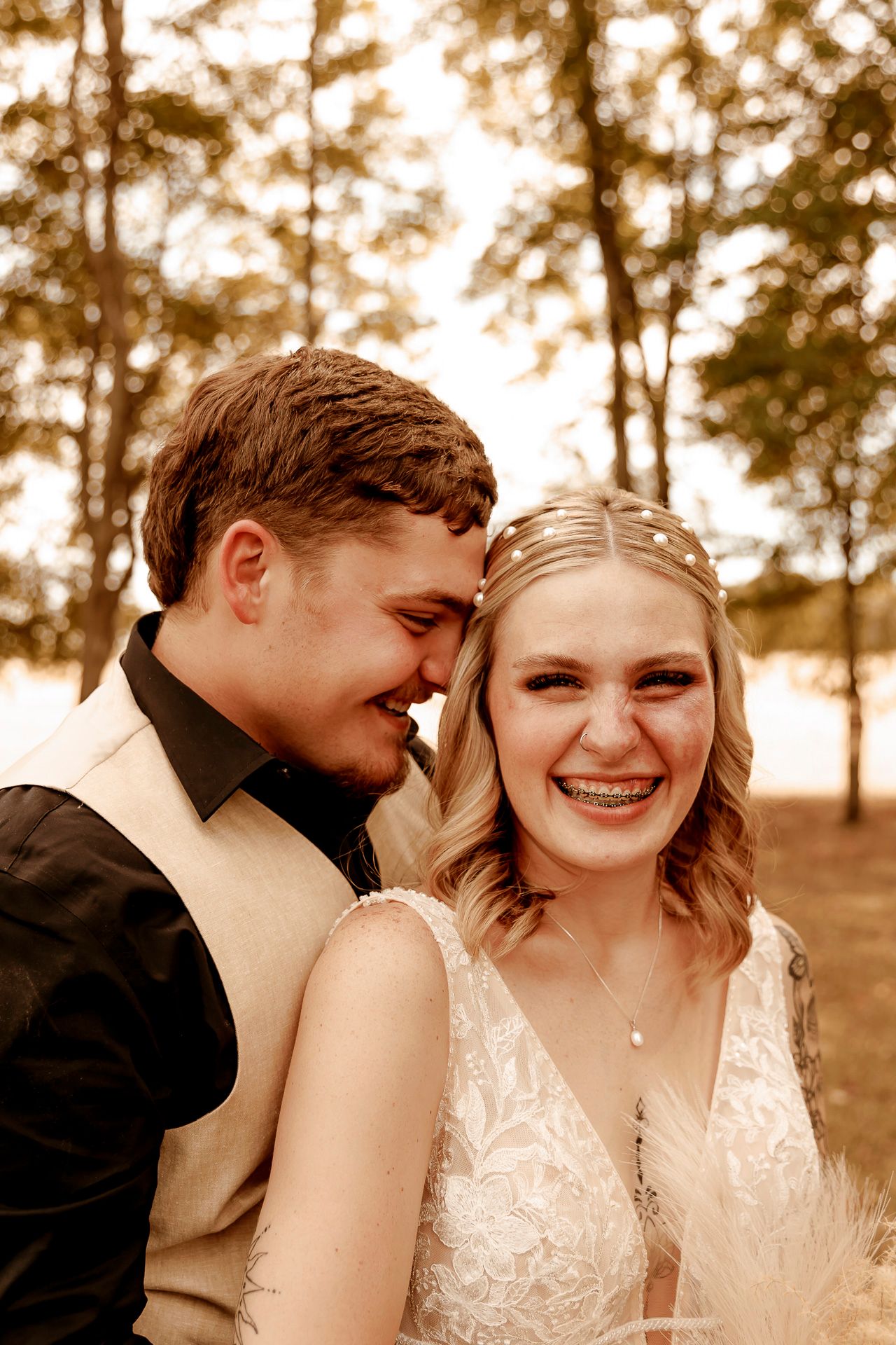 A joyful bride in lace dress and groom in tan vest share an intimate moment laughing together outdoors among trees.