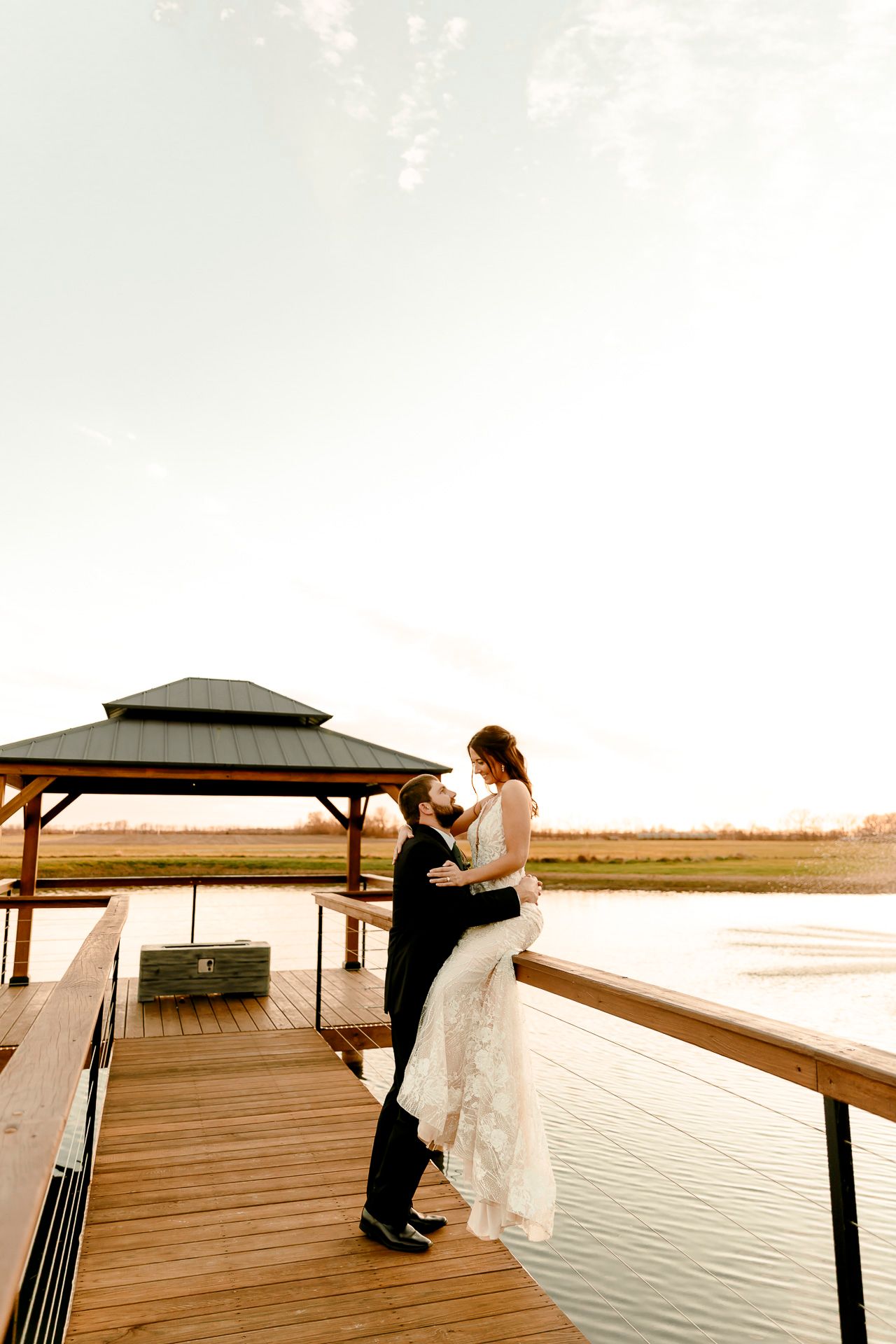 Groom lifting bride in white lace dress on wooden dock by lake at sunset near gazebo.