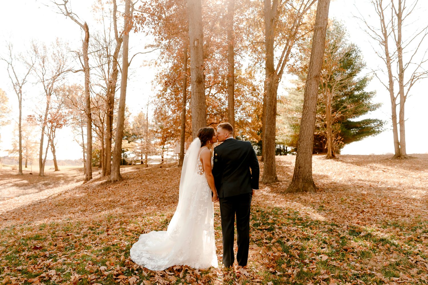 Bride and groom holding hands in a sunlit autumn forest, surrounded by fallen leaves and golden trees.
