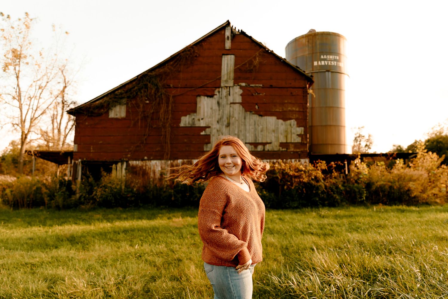 Young woman in orange sweater smiling in front of rustic red barn and silo at golden hour.