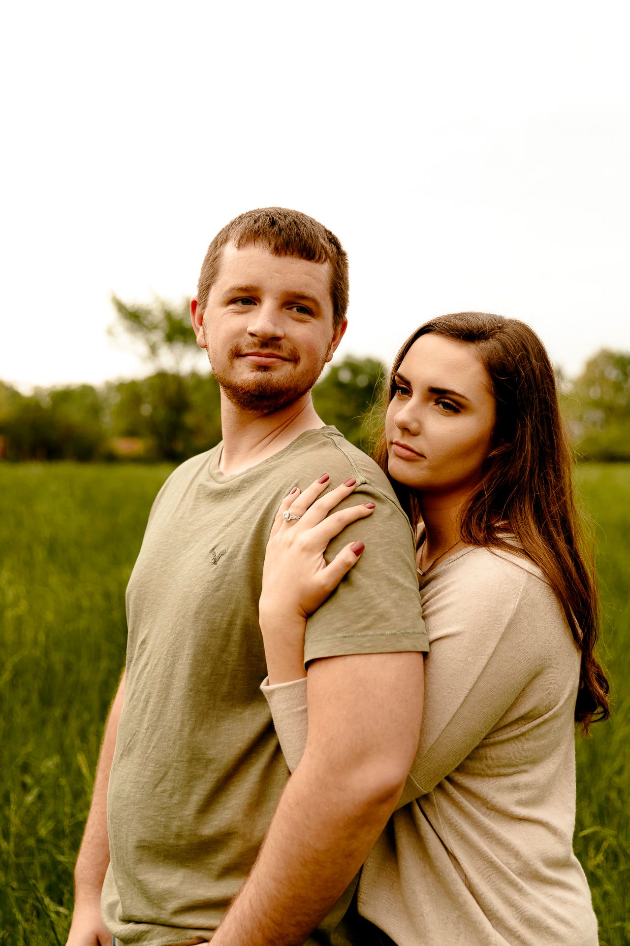 A young couple embraces in a green field, woman resting her chin on man's shoulder, both wearing olive toned shirts.