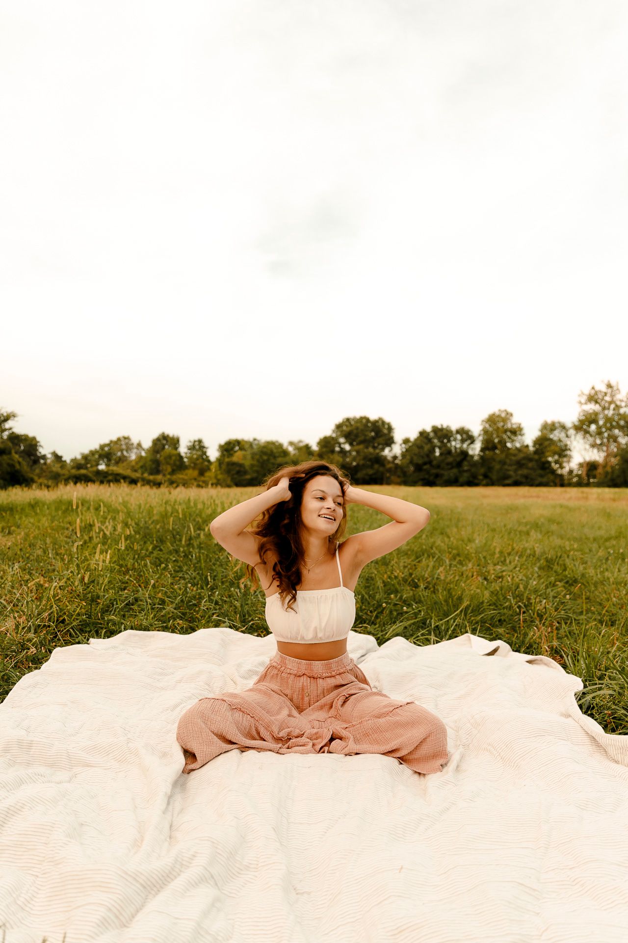 Young woman sitting cross-legged on white blanket in green field, smiling with hands in hair, wearing crop top and flowy pants.