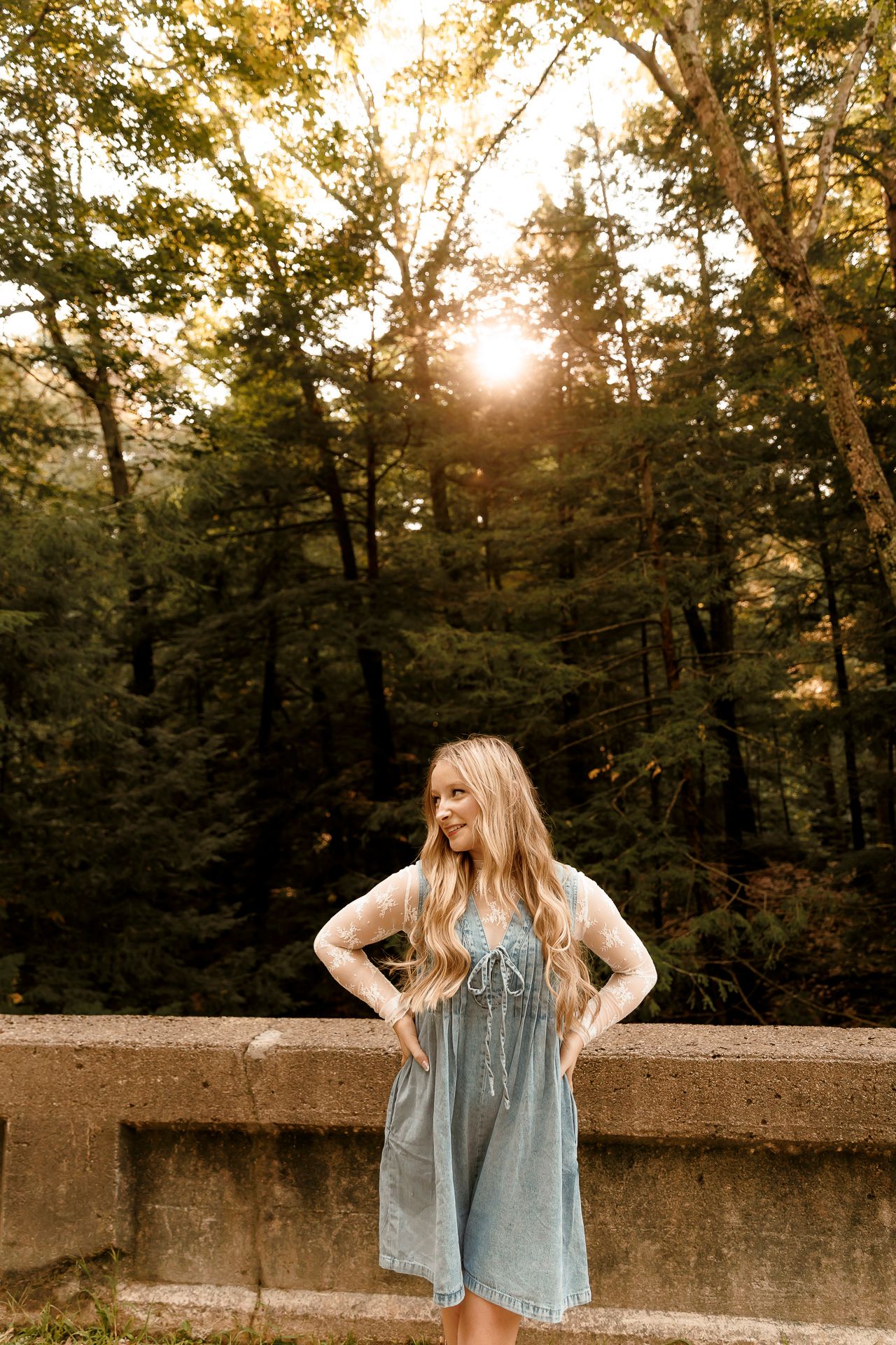 Young woman in light blue dress poses on stone bridge surrounded by lush green forest with golden sunset light.