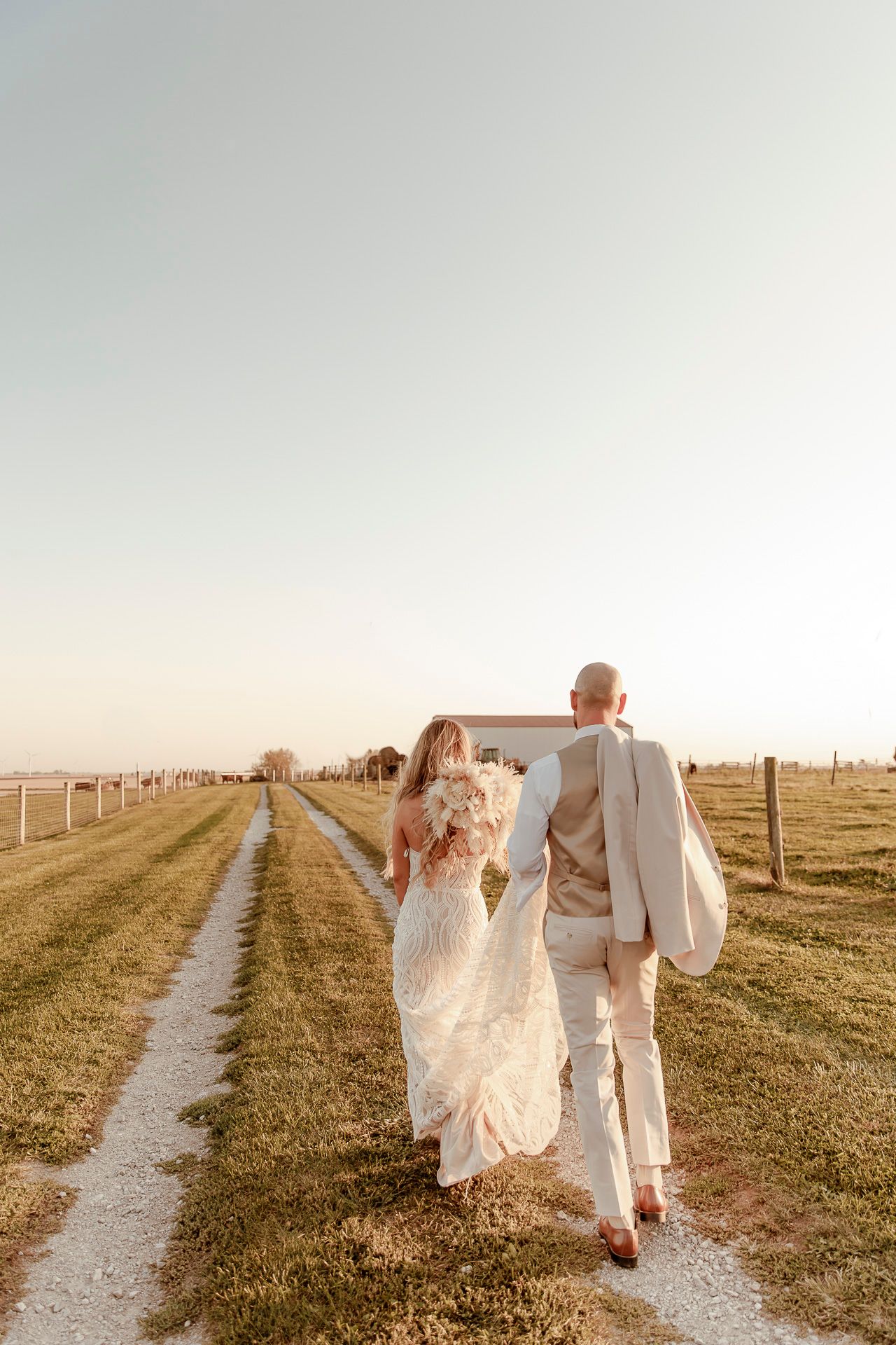 A couple walks hand in hand down a dirt path at sunset wearing elegant white wedding attire.