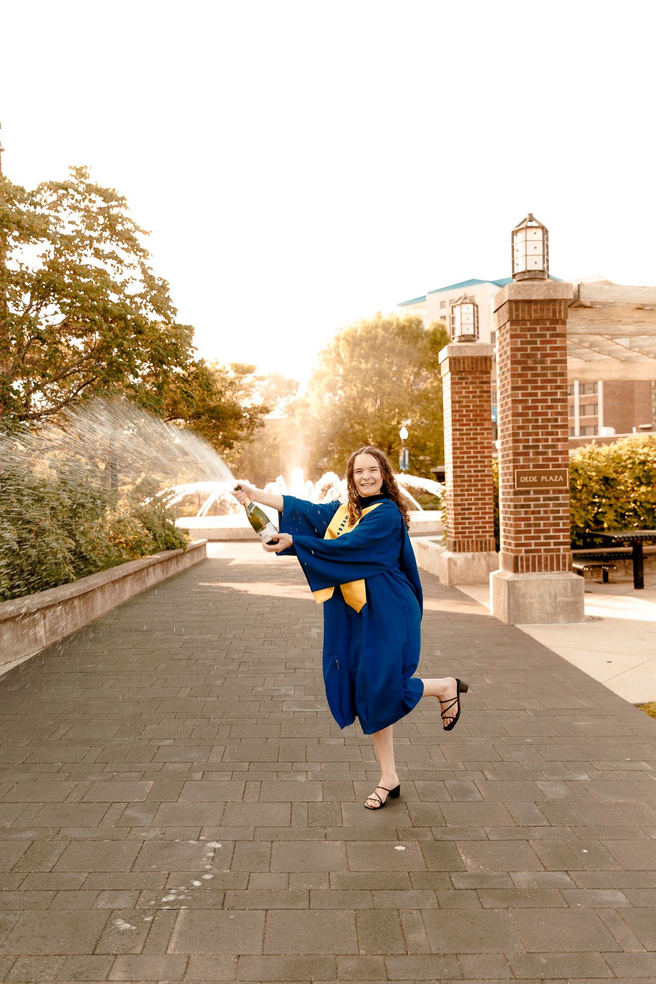 A joyful graduate in blue and gold cap and gown sprays champagne on a brick campus walkway with fountain backdrop.