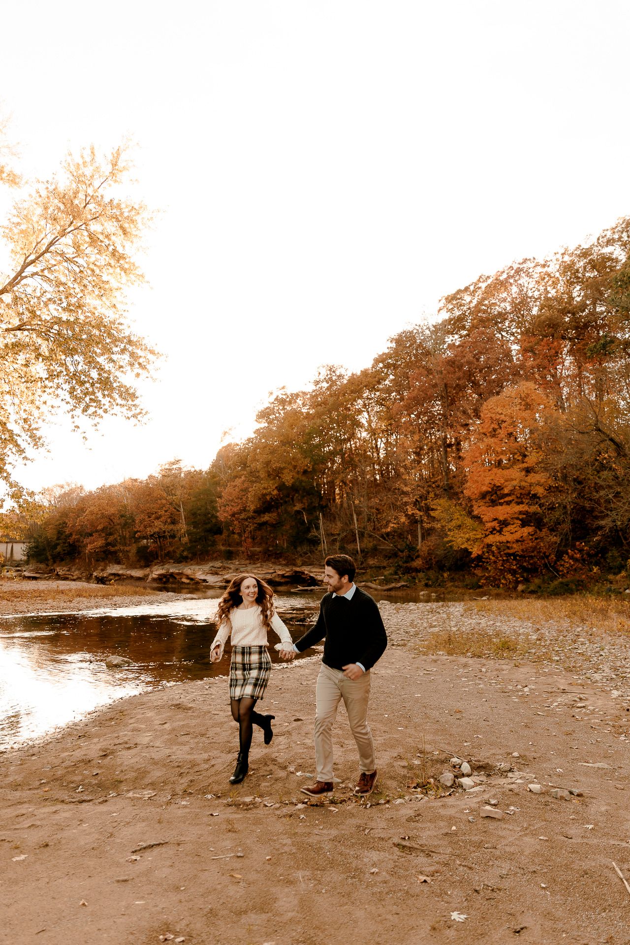 A couple holds hands and runs along a sandy riverbank surrounded by vibrant autumn foliage in warm orange and red tones.