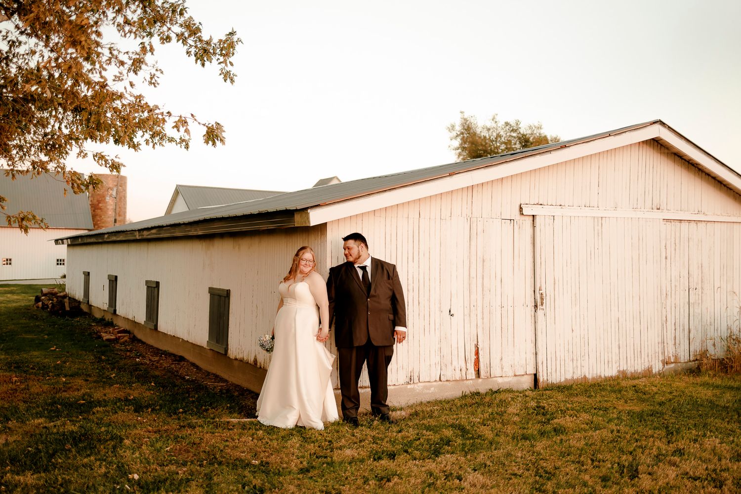 Bride and groom holding hands walking in front of a rustic white barn on their wedding day.