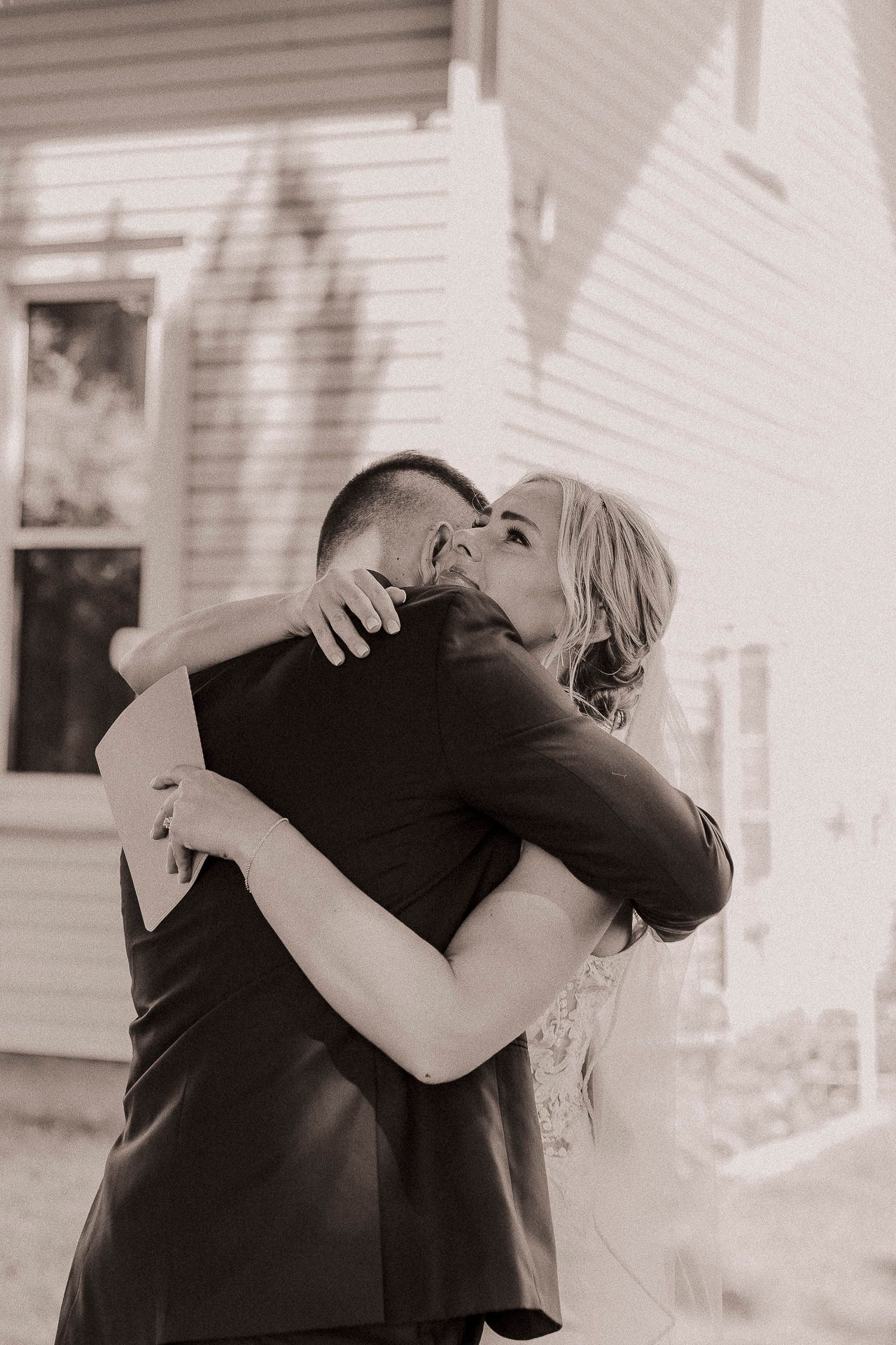 Black and white photo of a bride and groom sharing an emotional embrace outside a white building on their wedding day.