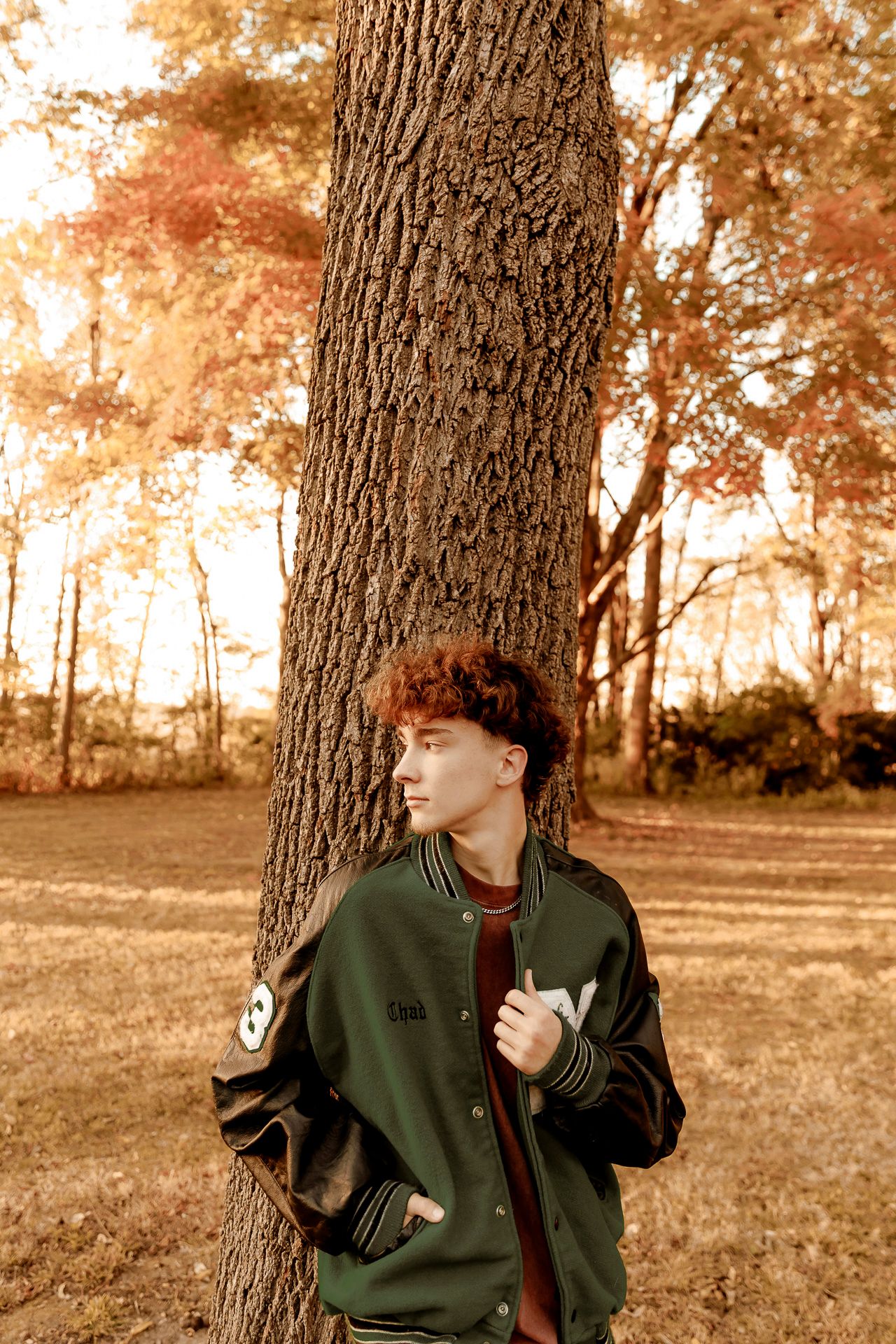 A figure in a dark green jacket poses against a tree trunk in an autumn setting with sepia-toned lighting.