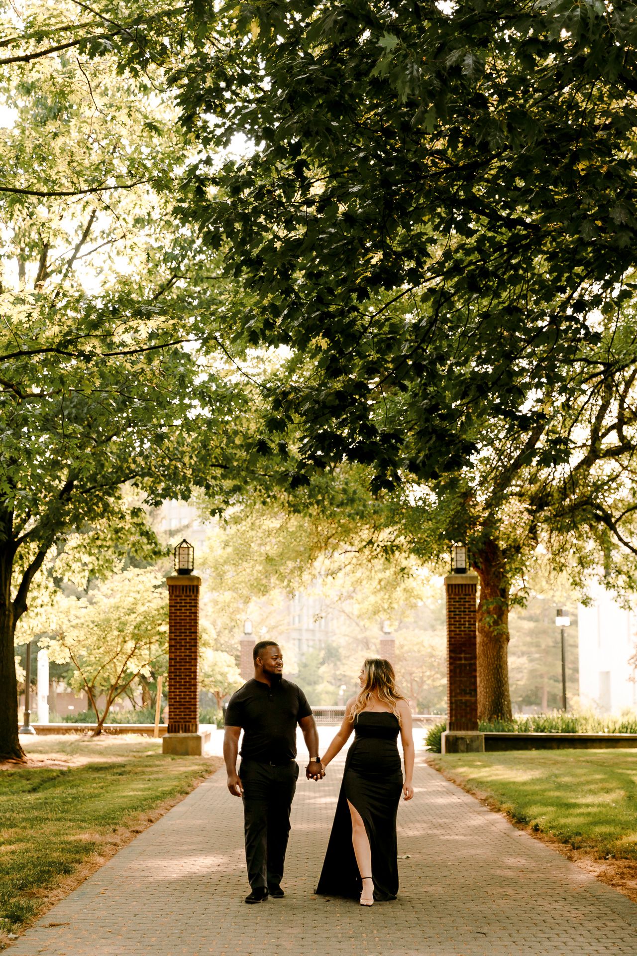 Couple in black formal attire holding hands on a tree-lined brick pathway at golden hour.