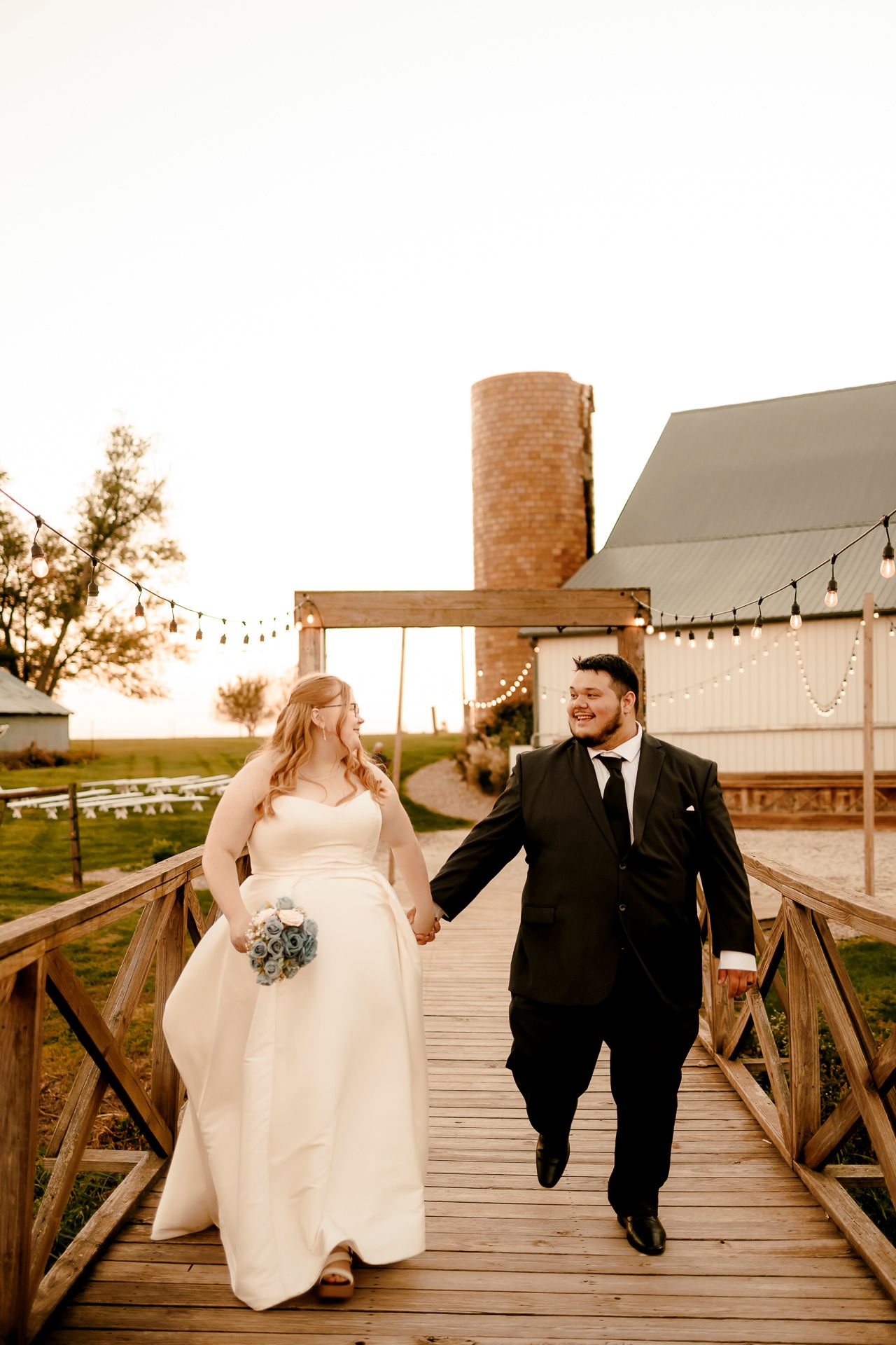Bride and groom hold hands walking on wooden bridge at golden hour near rustic barn wedding venue.
