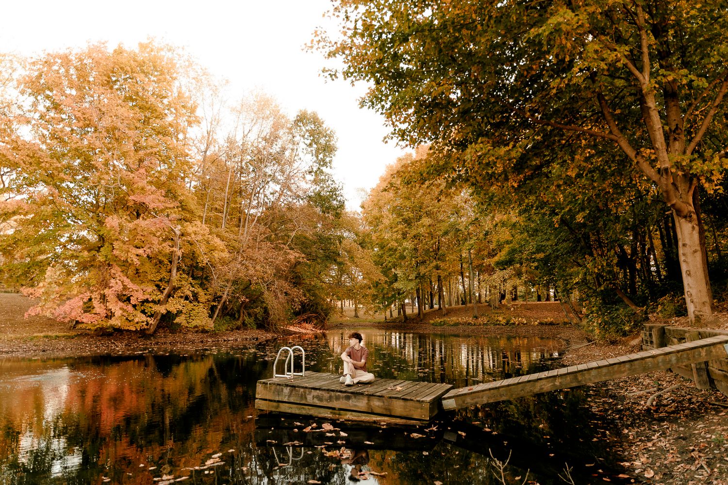 Person sitting on a wooden dock surrounded by autumn trees reflecting on a calm pond in fall.