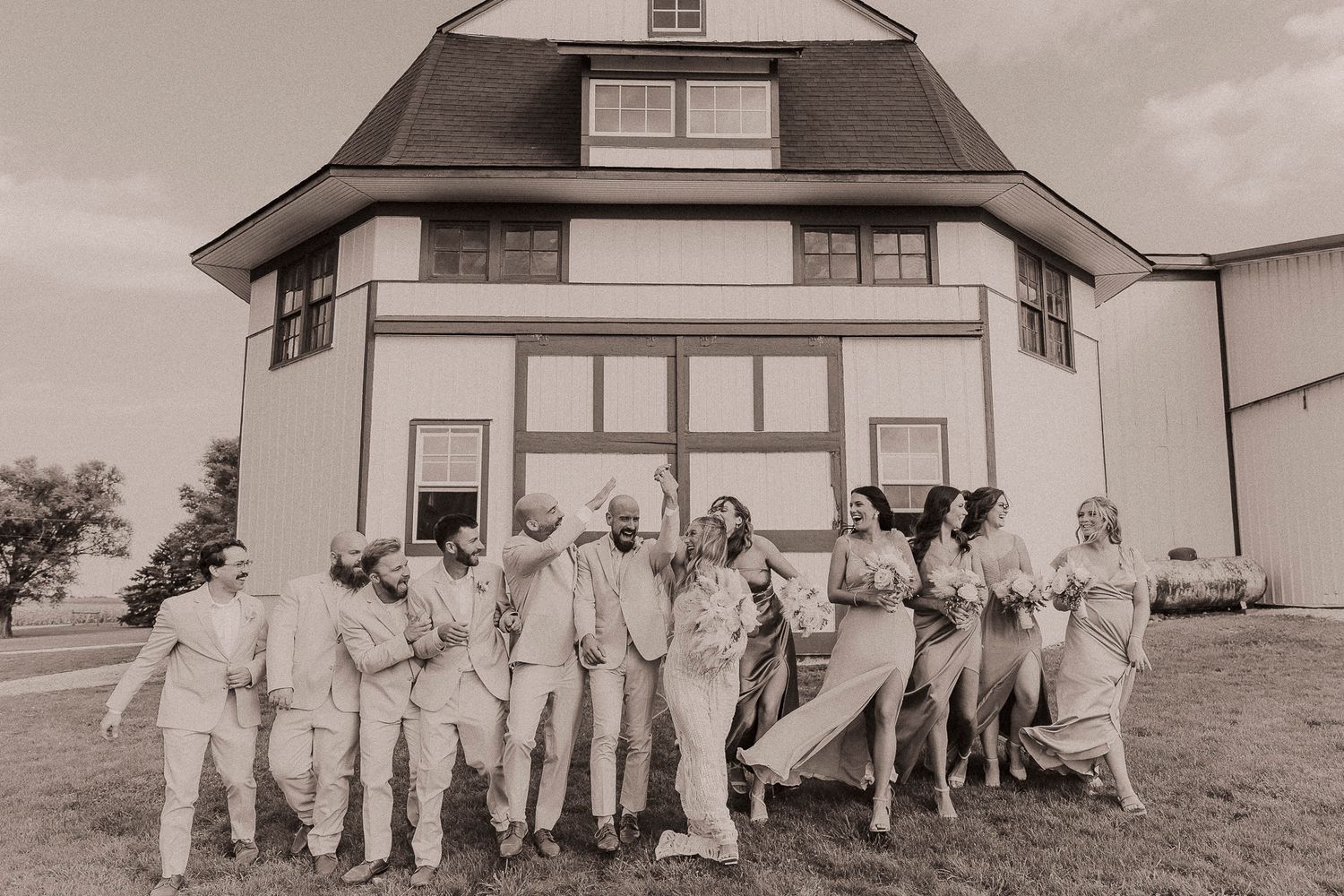 Wedding party posing and laughing in front of a white barn venue in a sepia-toned outdoor group photo.