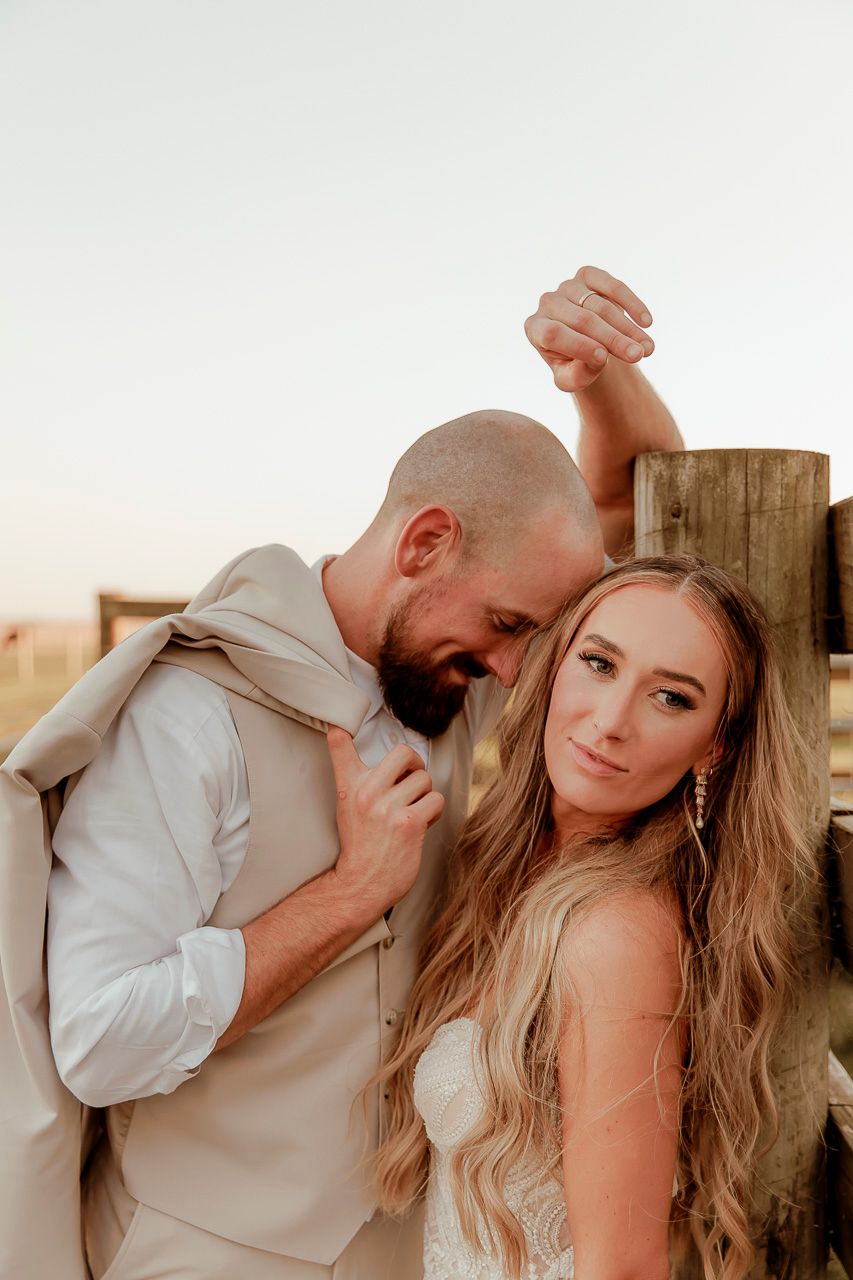 A romantic sunset portrait of a couple in wedding attire leaning against a wooden fence post during golden hour.